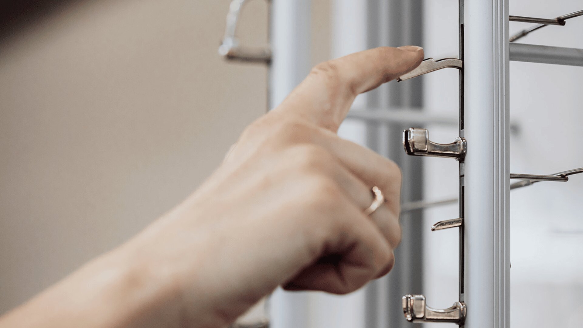 A close-up of a hand adjusting horizontal metal blinds on a window, with fingers gripping one of the slats. The background is softly blurred.