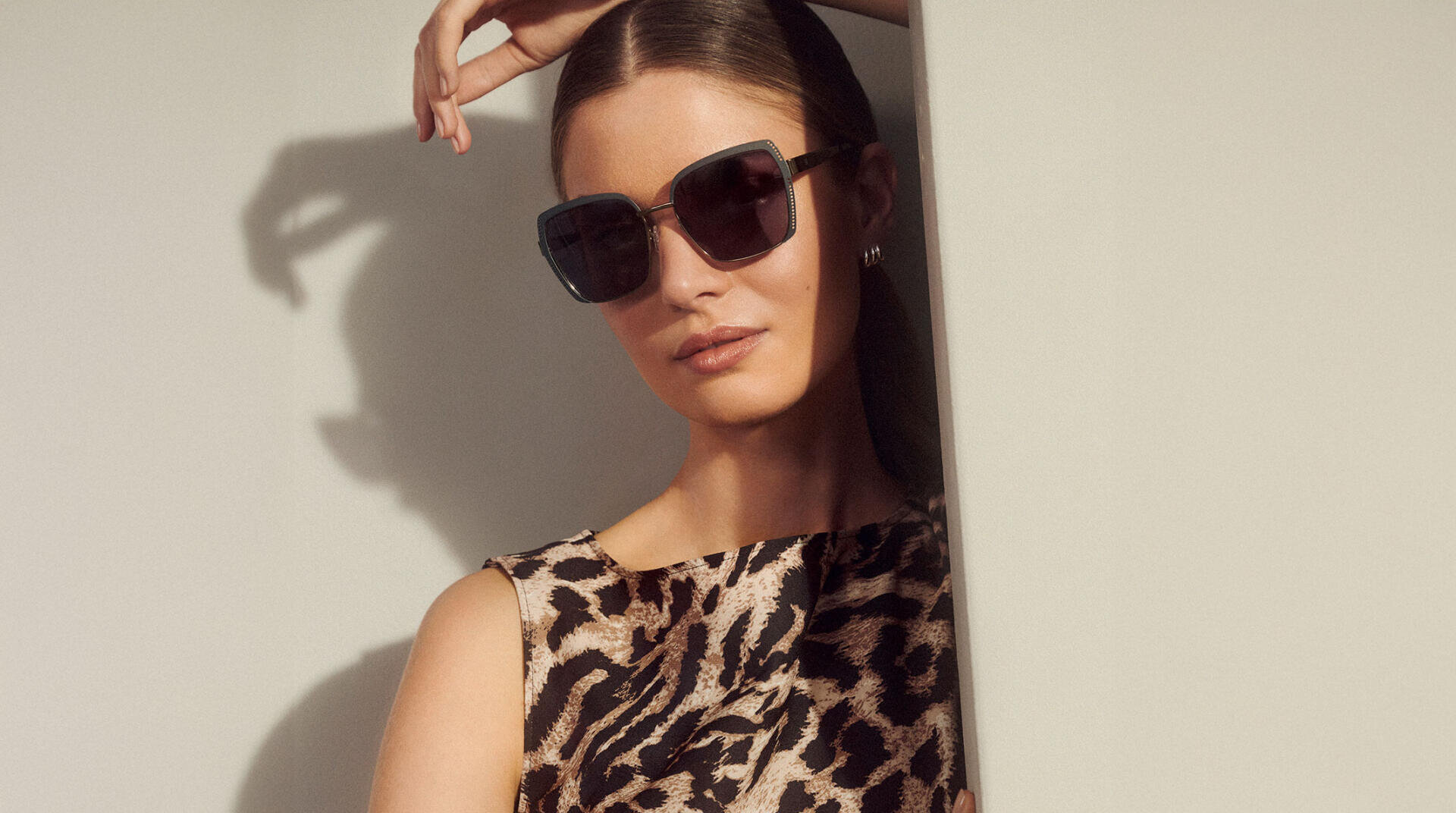 A woman wearing large black sunglasses and a sleeveless leopard-print top is leaning against a light-colored wall, her hair pulled back and one hand over her head.