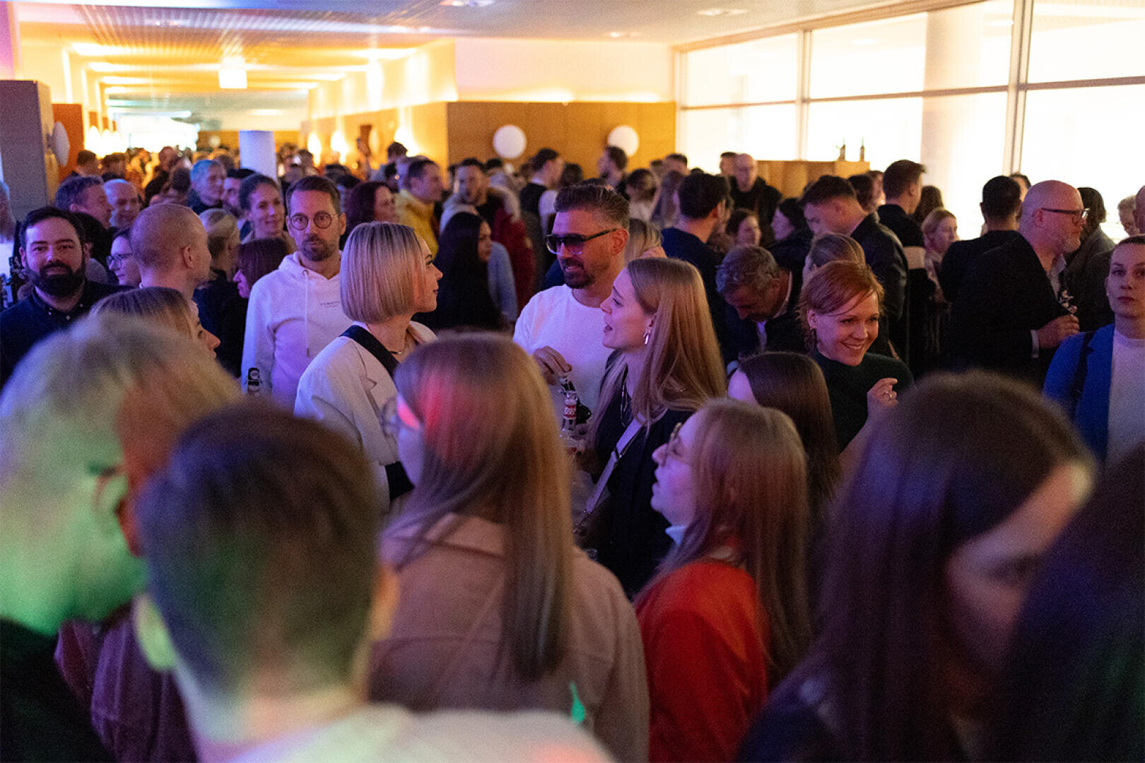 A large group of people gather in a brightly lit hallway during an event, many are chatting and some are holding drinks in their hands. The atmosphere is lively and crowded.