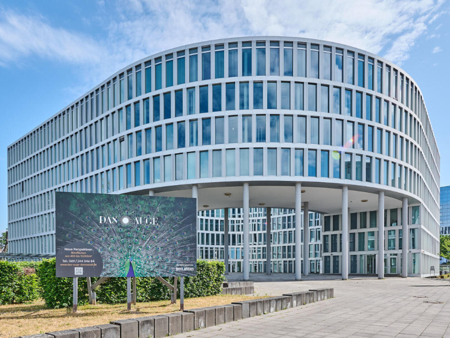 Ein modernes, geschwungenes Bürogebäude mit vielen Fenstern steht hinter einem Schild mit der Aufschrift "DAX Allee" und einer abstrakten Grafik. Der Himmel ist teilweise bewölkt und die Fläche ist gepflastert mit etwas Grün davor.