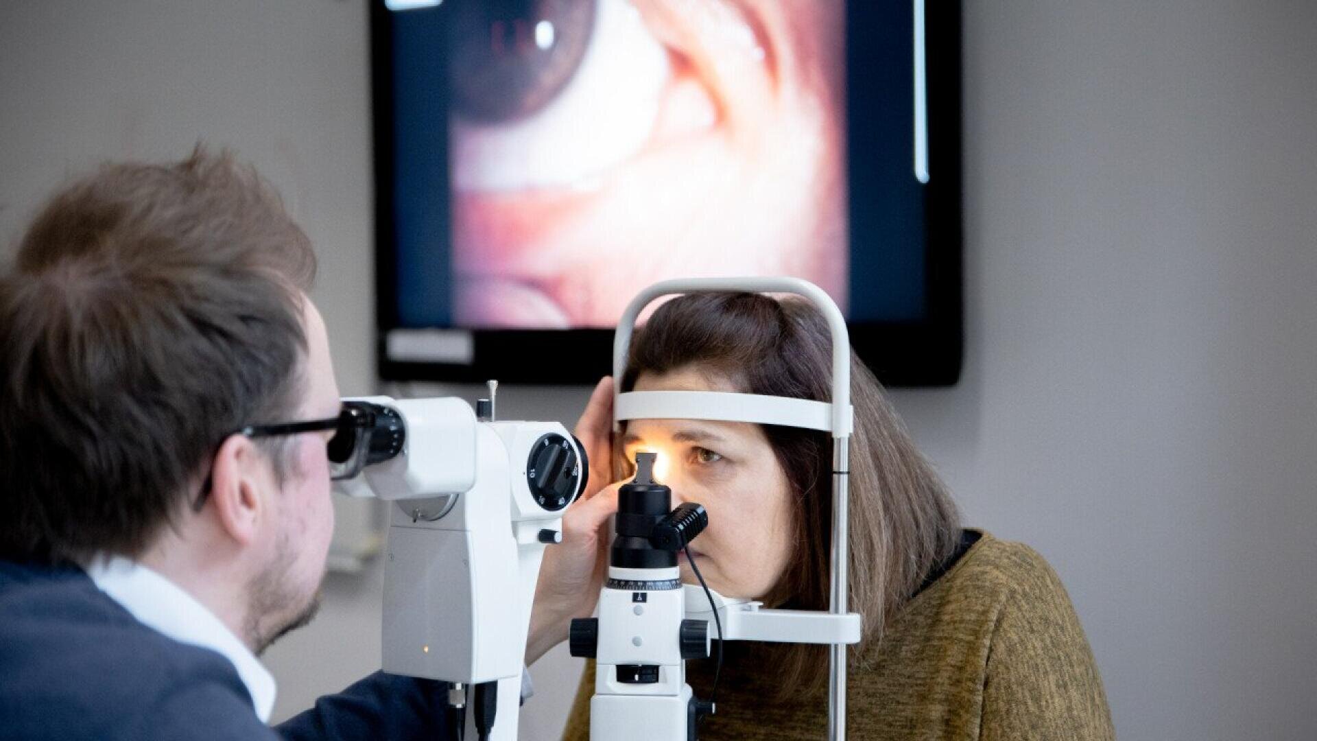 An eye doctor examines a womans eye with a slit lamp while an enlarged image of an eye is displayed on a screen in the background.
