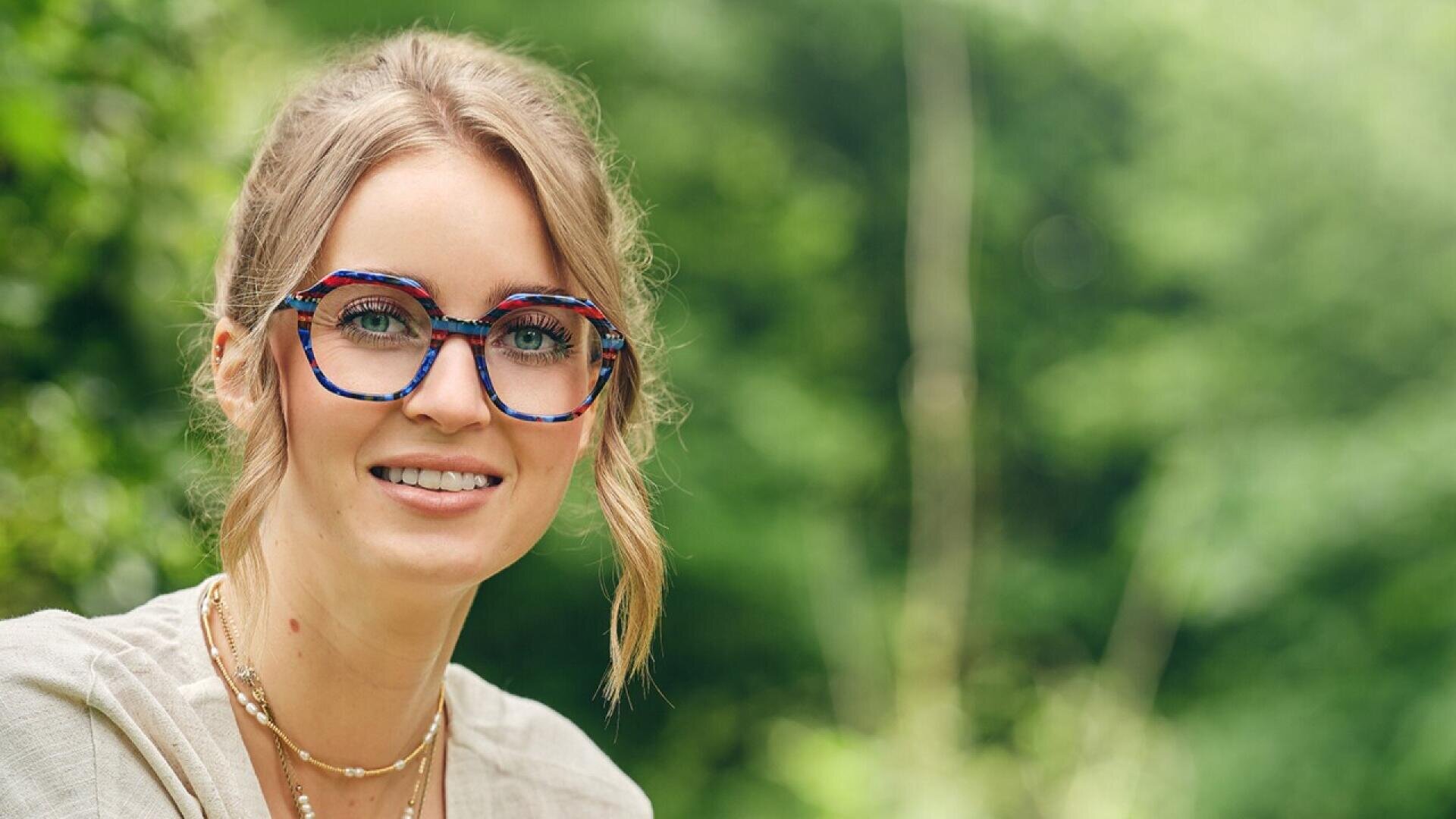 A woman with blonde hair in a loose updo, wearing colorful round glasses and layered necklaces, smiles outdoors in front of a blurred green, leafy background.