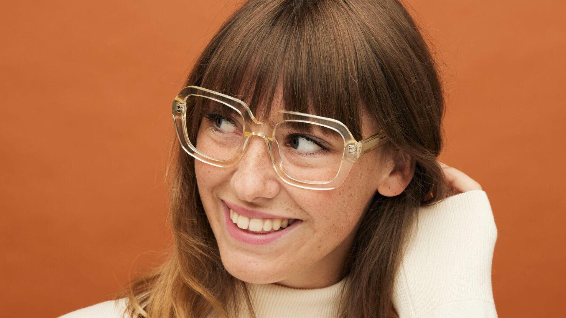A smiling woman with long brown hair and bangs wears oversized clear glasses and a cream-colored turtleneck sweater, standing in front of an orange background.
