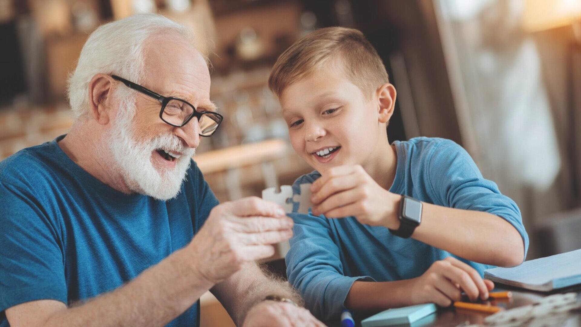 An elderly man and a young boy smile and work together on a puzzle at a table, both wearing blue shirts. The scene is warm and bright, suggesting a joyful moment of bonding.