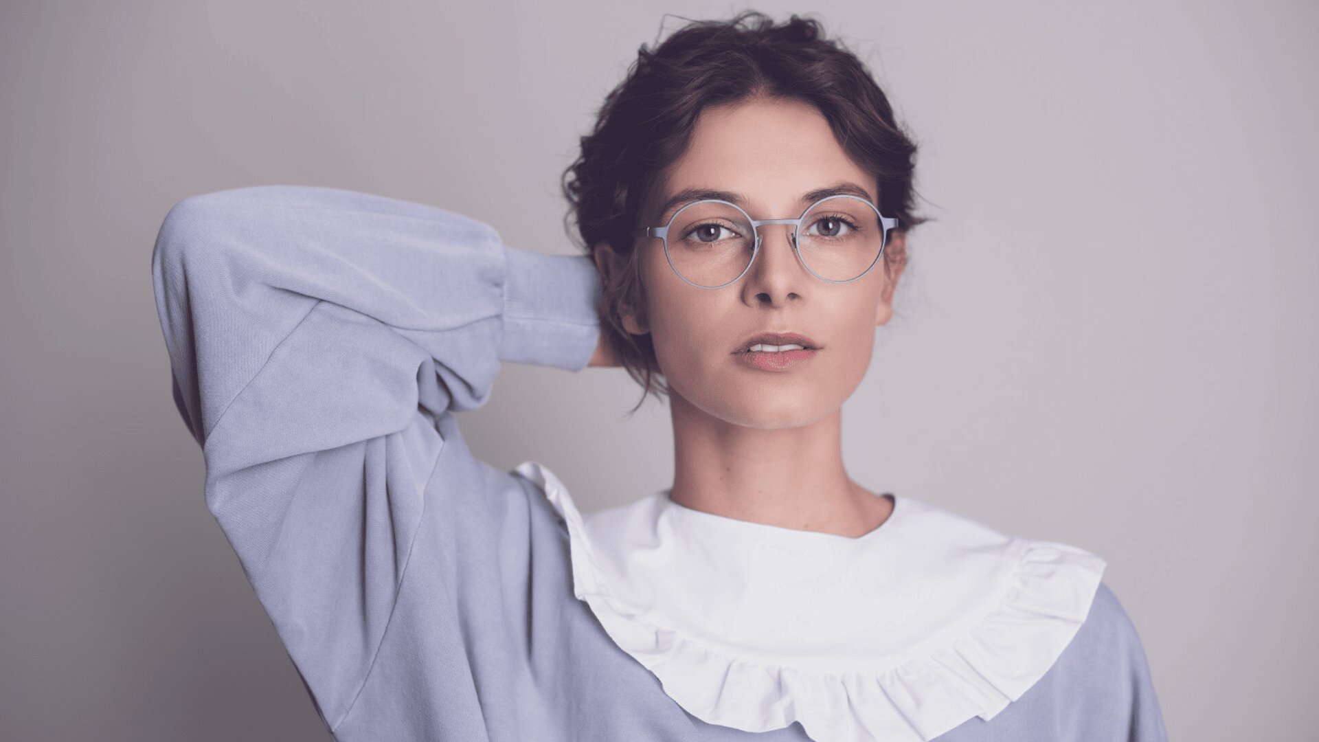 Eine Person mit kurzen, gelockten dunklen Haaren trägt eine Brille und ein hellviolettes Oberteil mit weißem Rüschenkragen und hält eine Hand hinter dem Kopf vor einem schlichten hellen Hintergrund.