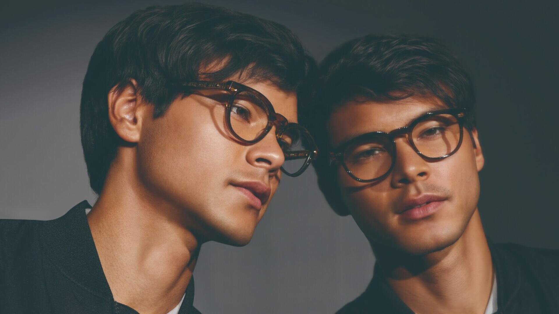 A young man wearing glasses looks at his own reflection in a mirror, creating the appearance of two similar faces side by side.