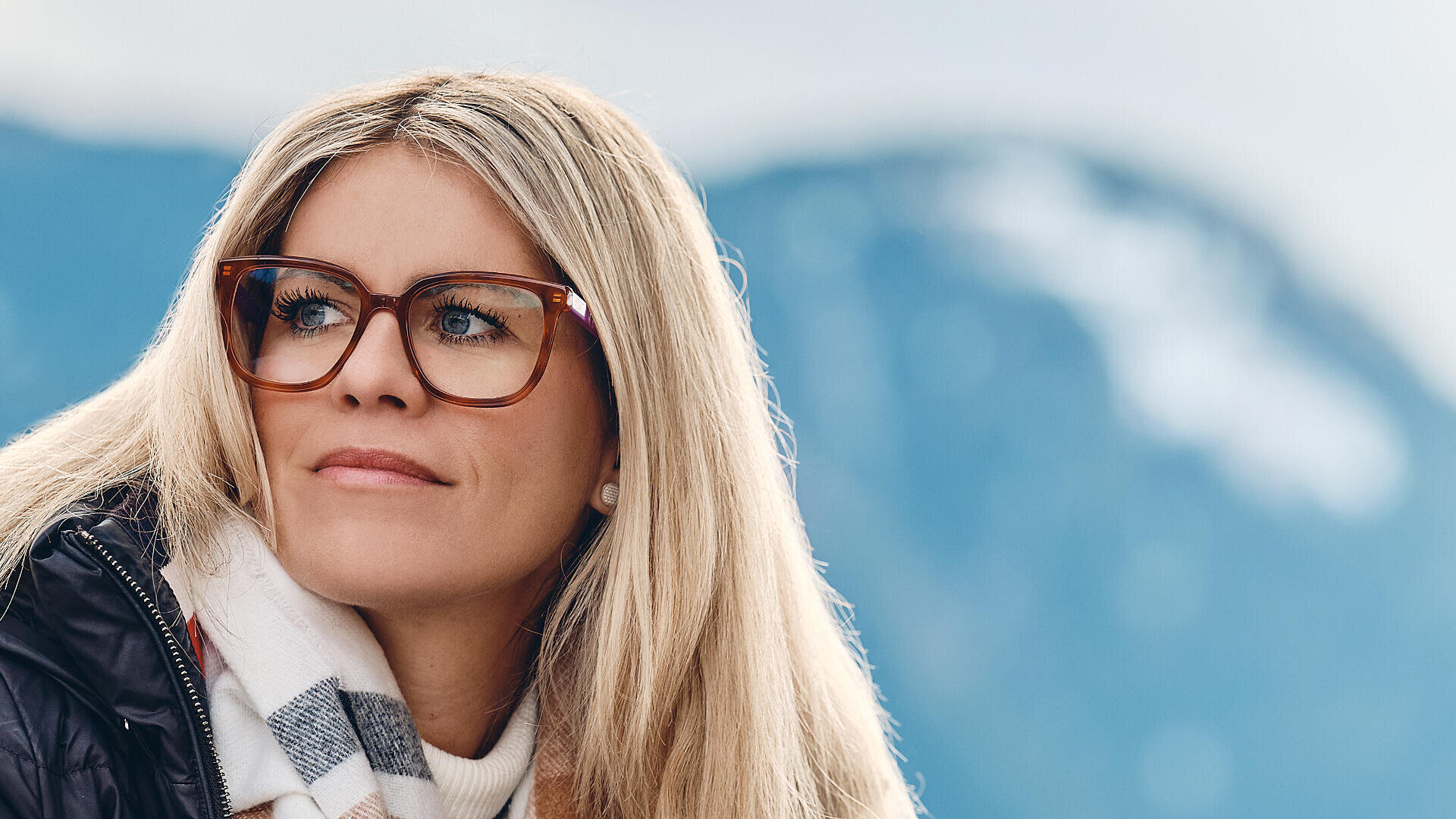 Eine Frau mit langen blonden Haaren und Brille blickt zur Seite, bekleidet mit Schal und Jacke, im Hintergrund ist eine verschwommene Berglandschaft zu sehen.