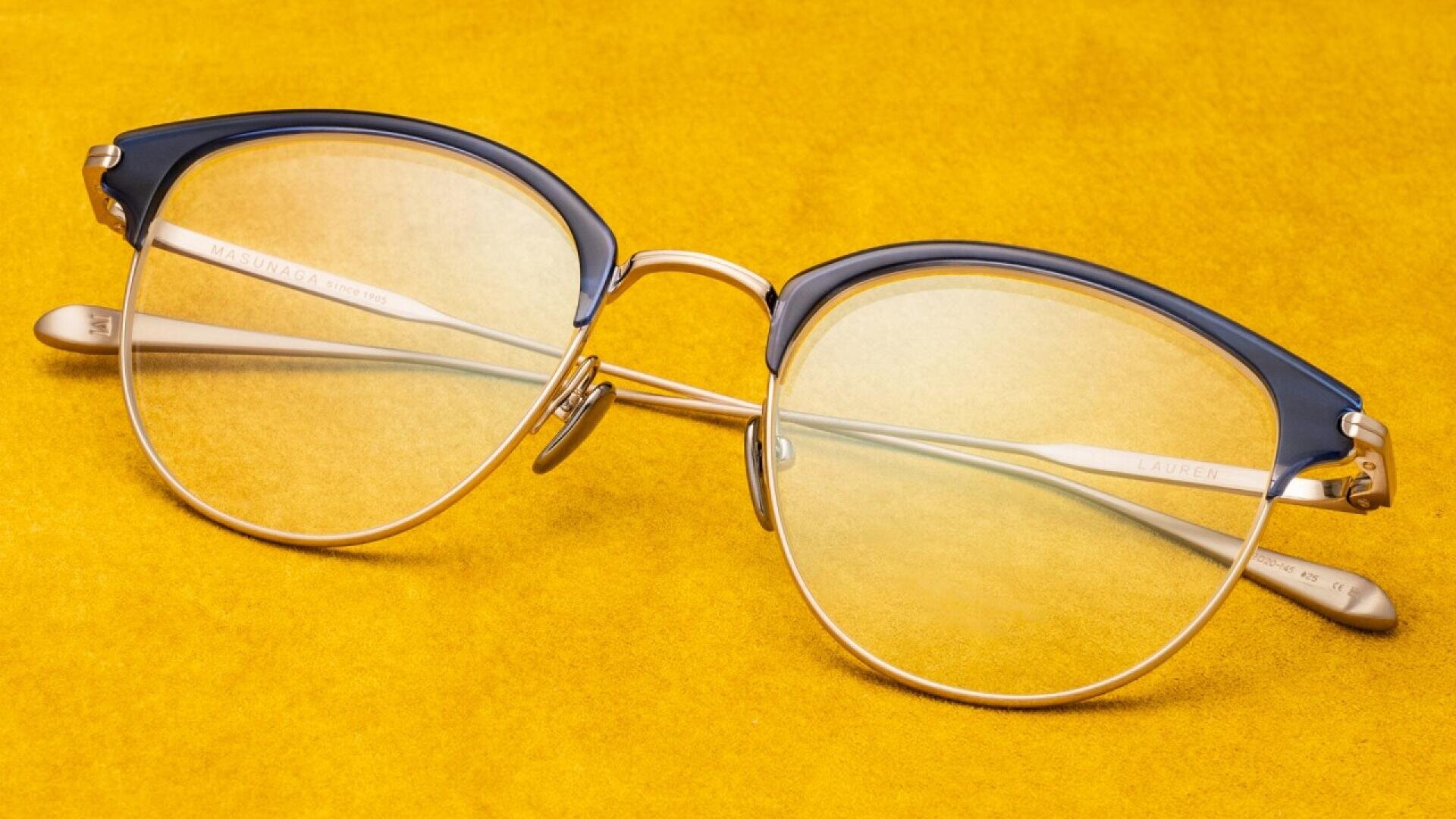 A pair of vintage-style eyeglasses with round gold frames and dark blue upper rims, placed on a bright yellow surface.