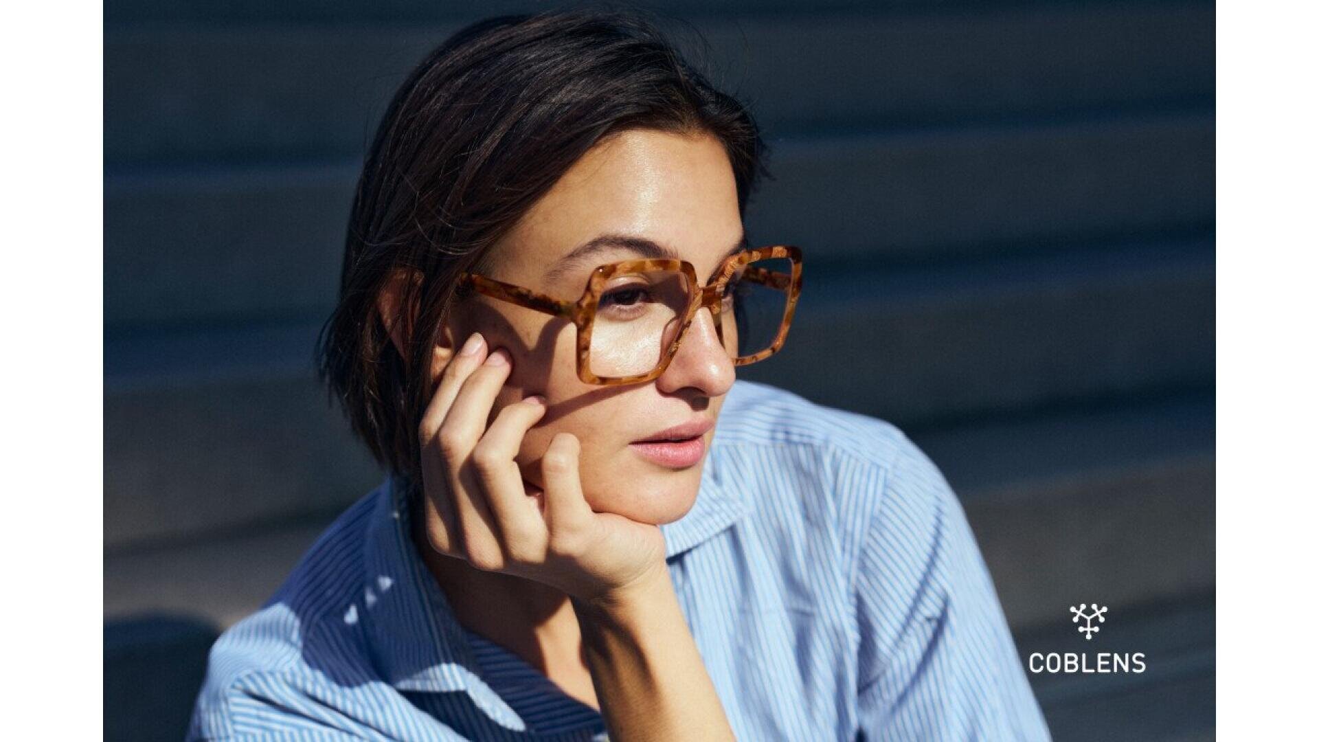 A person wearing oversized, square tortoiseshell glasses and a blue striped shirt rests their chin on their hand. The background is blurred, showing steps. The COBLENS logo appears in the bottom right corner.