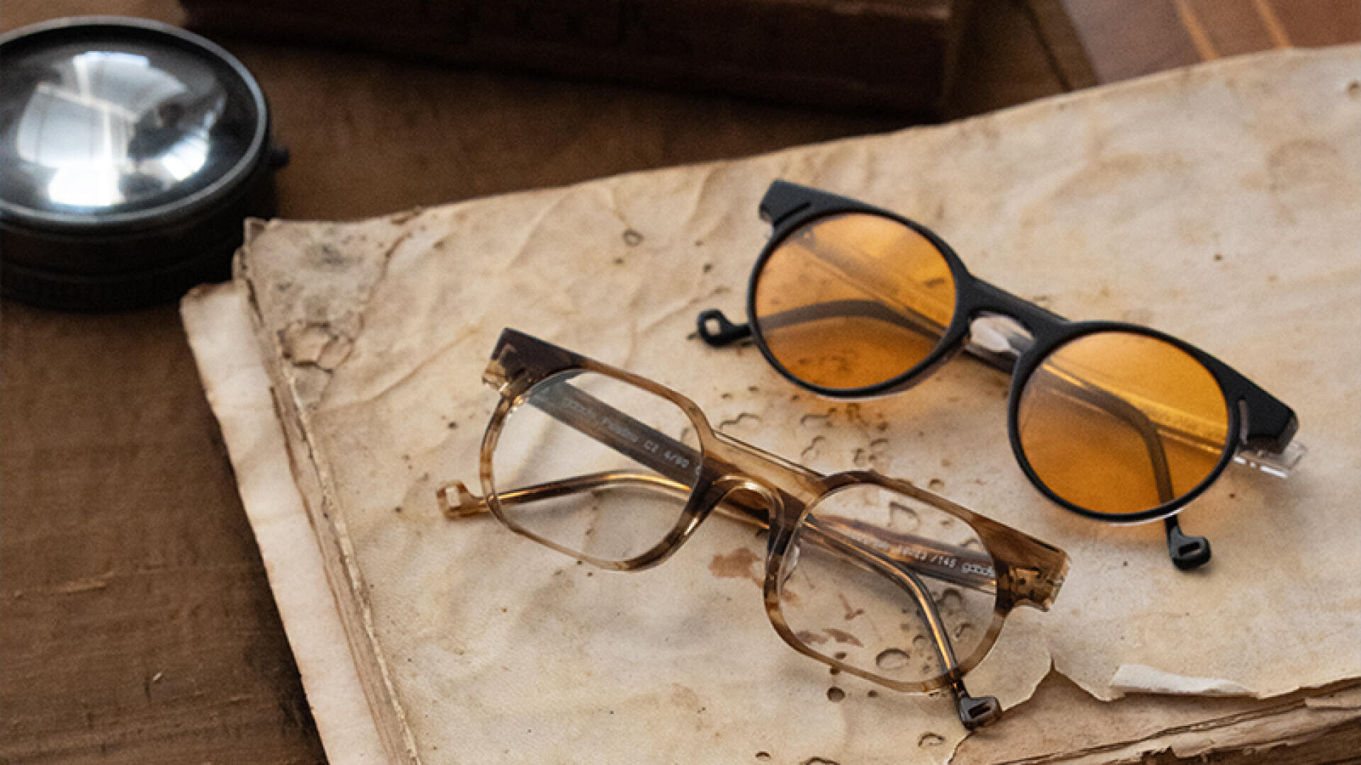 Zwei Brillen, eine mit klaren Gläsern und braunem Rahmen, die andere mit runden, bernsteinfarbenen Gläsern und schwarzem Rahmen, liegen auf einem abgenutzten, vergilbten Buch neben einer großen Lupe.
