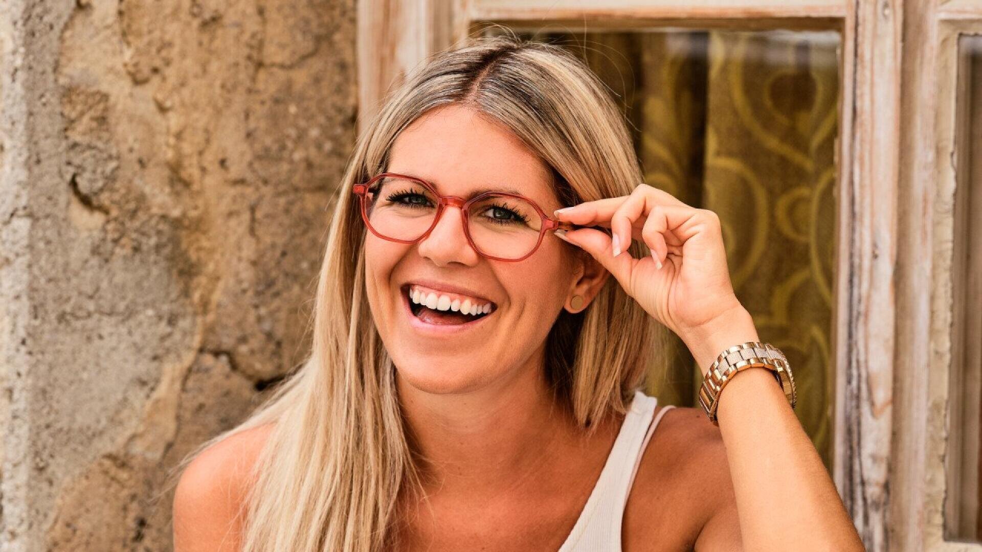 A smiling woman with long blonde hair and red-framed glasses sits in front of a rustic wall and window, wearing a white sleeveless top and a gold watch.