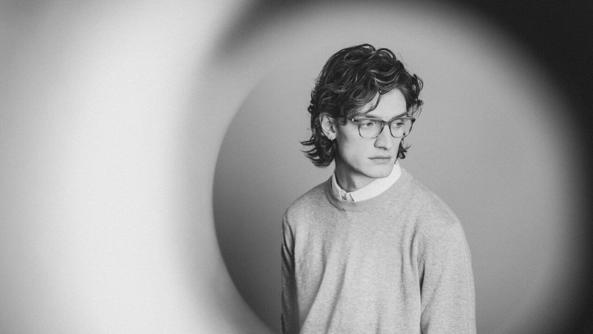 A young person with wavy hair and glasses, wearing a light sweater over a collared shirt, looks to the side. The black and white photo is framed by a blurred circular shape in the foreground.
