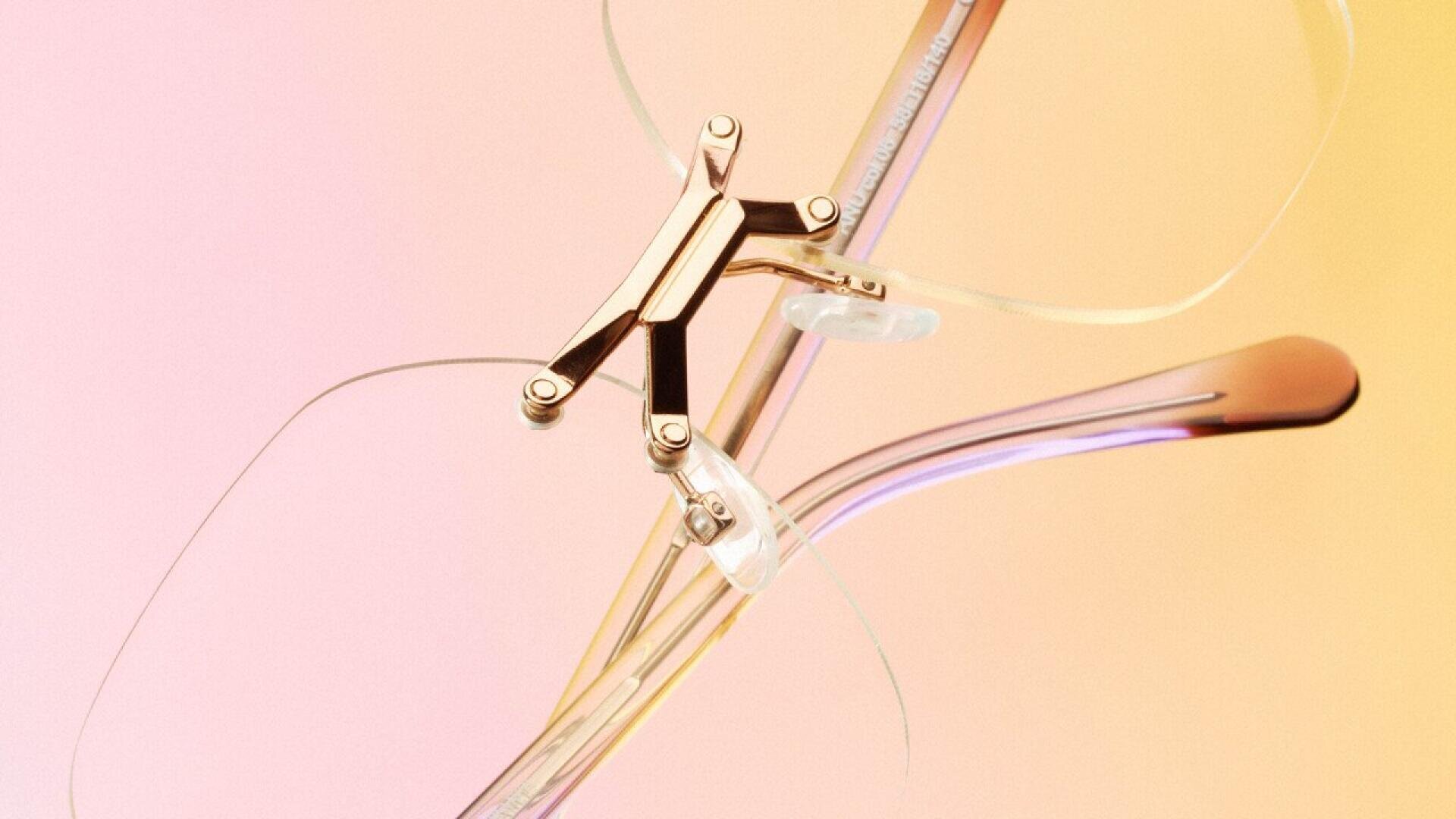 Close-up of stylish, rimless eyeglasses with a metallic bridge and transparent nose pads, set against a softly blended pink and yellow gradient background.