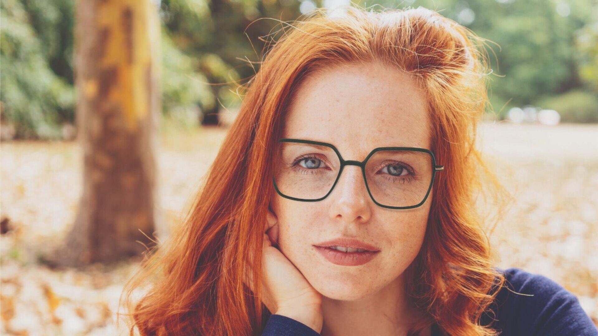 A woman with long red hair and green glasses looks directly at the camera, resting her chin on her hand. She is outdoors with trees and fallen leaves in the background.
