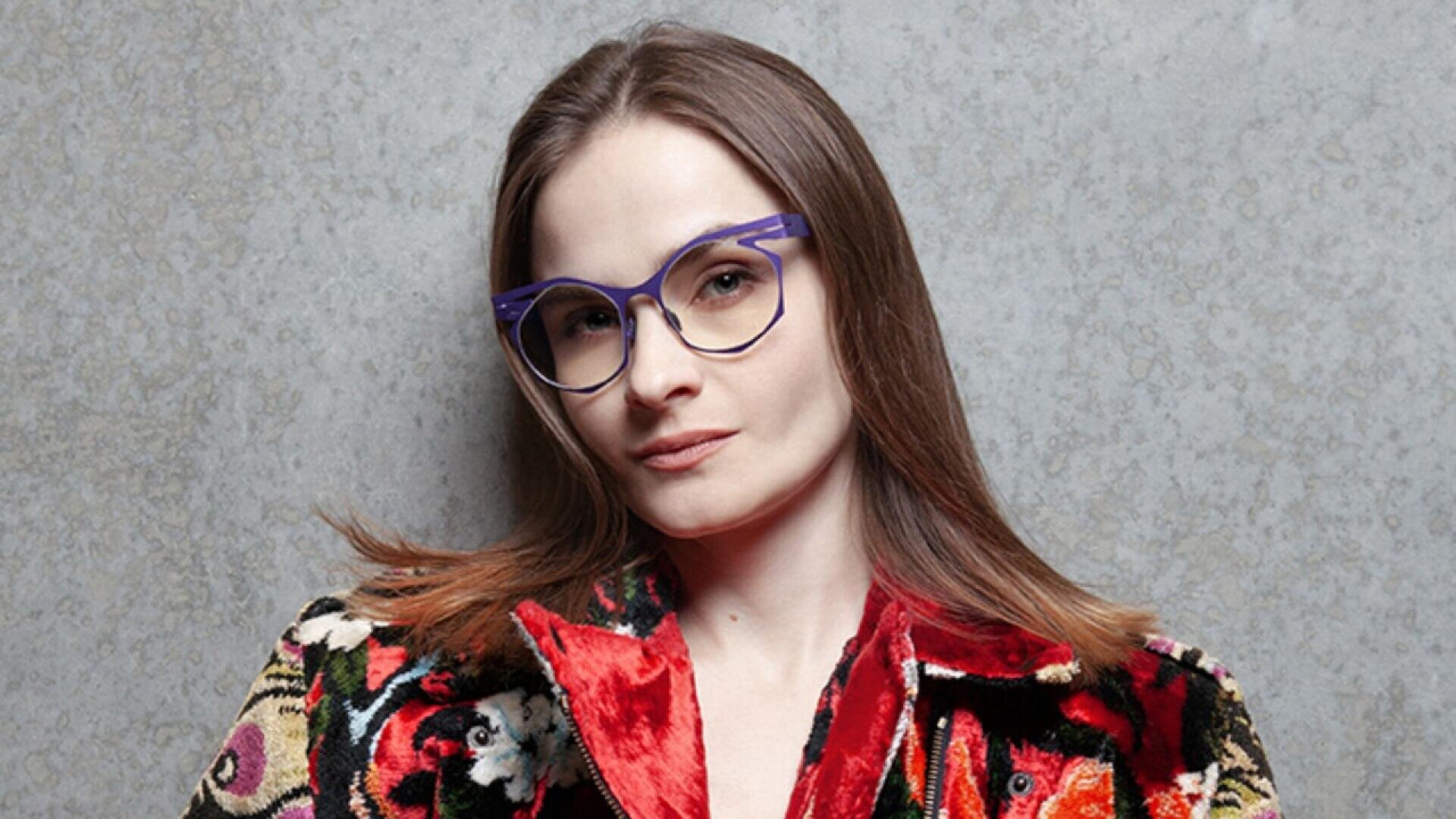 A woman with straight, shoulder-length brown hair and purple glasses stands in front of a gray textured wall, wearing a colorful, patterned velvet jacket.