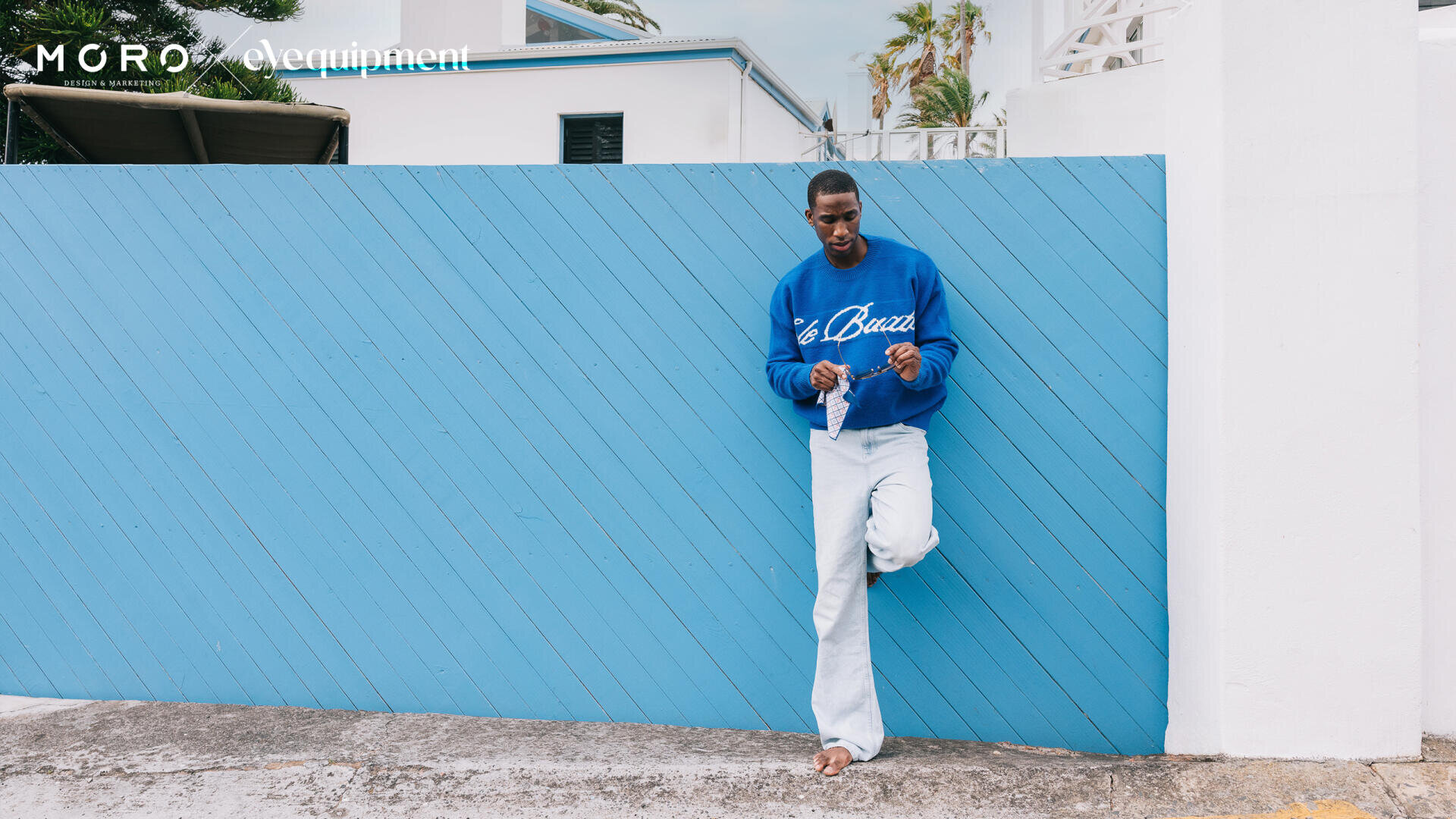 Ein Mann in einem blauen Sweatshirt und hellen Jeans lehnt lässig an einem schrägen blauen Holzzaun und hält eine Sonnenbrille in der Hand. Im Hintergrund sind weiße Gebäude und Grünflächen zu sehen.