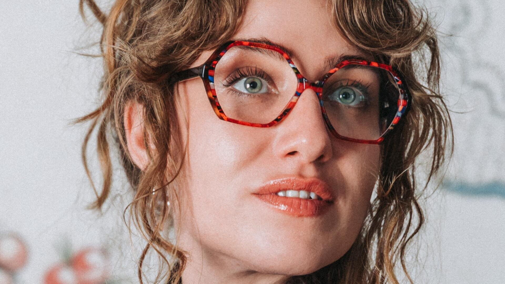 Eine Frau mit gewelltem hellbraunem Haar und einer großen, bunten, runden Brille blickt leicht nach oben und lächelt sanft. Der Hintergrund ist unscharf.