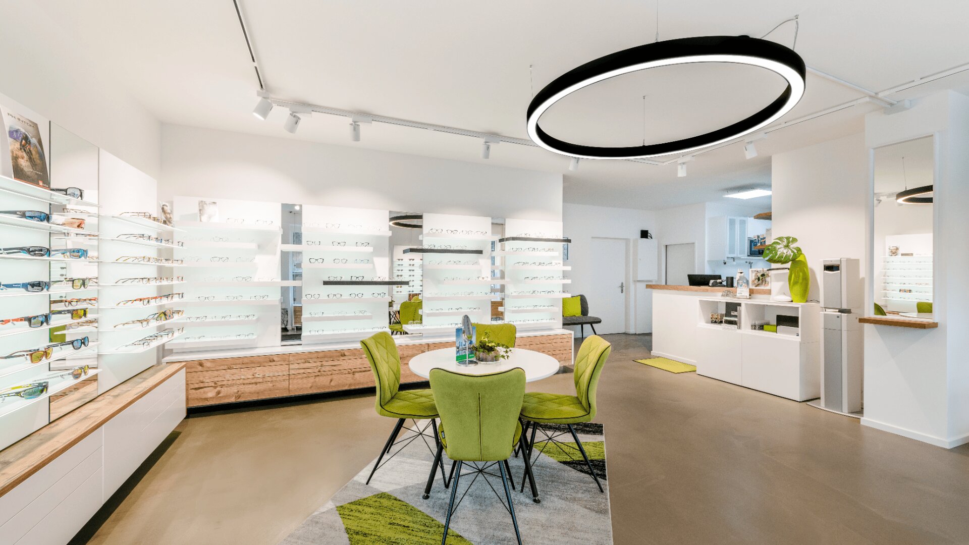 Modernes Optikergeschäft mit heller Beleuchtung, grünen Stühlen um einen runden Tisch, Ausstellungswänden mit Brillengestellen und einem weißen Tresen mit grünen Akzenten in einem sauberen, modernen Ambiente.