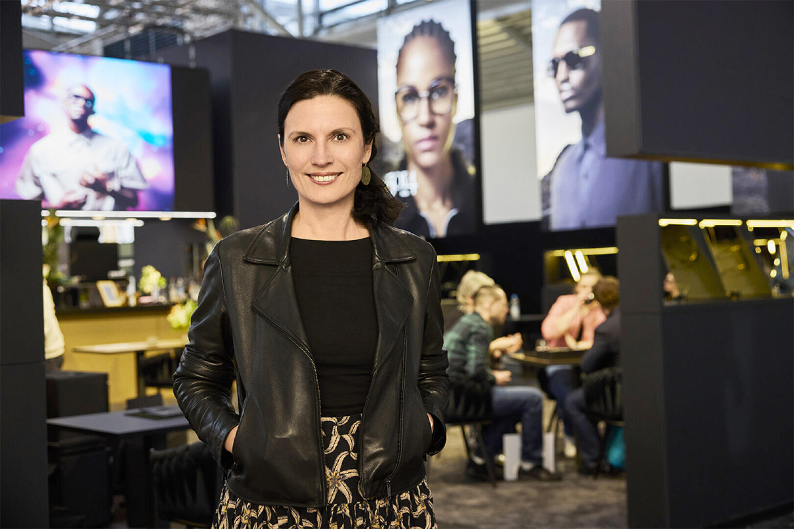 A woman in a black leather jacket stands smiling in a modern office with people working in the background and large portraits hanging on the walls.