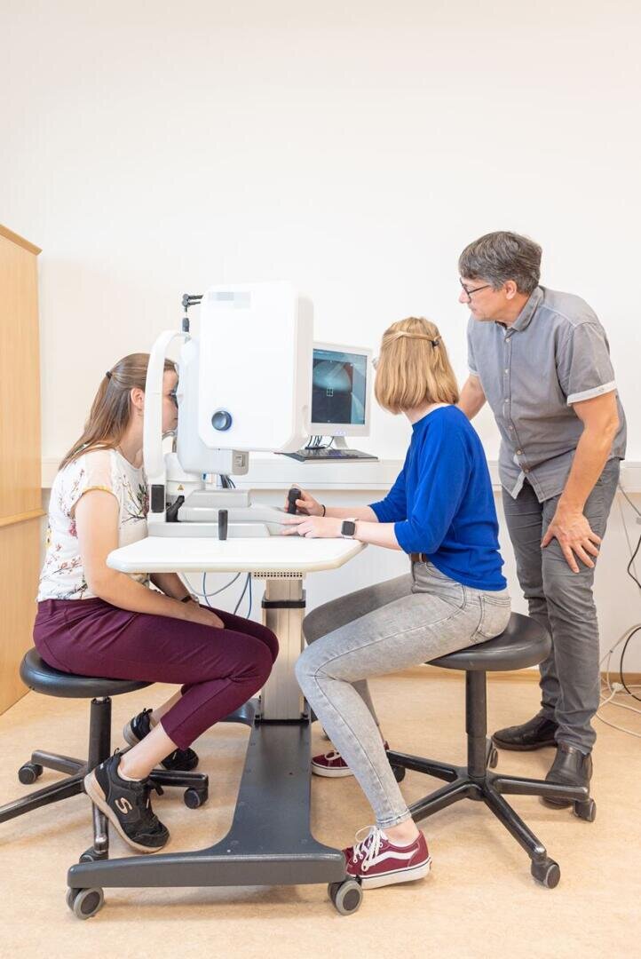 Two women sit at an ophthalmology machine, one operates the controls, the other has her eyes examined, while a man stands nearby and observes and assists.