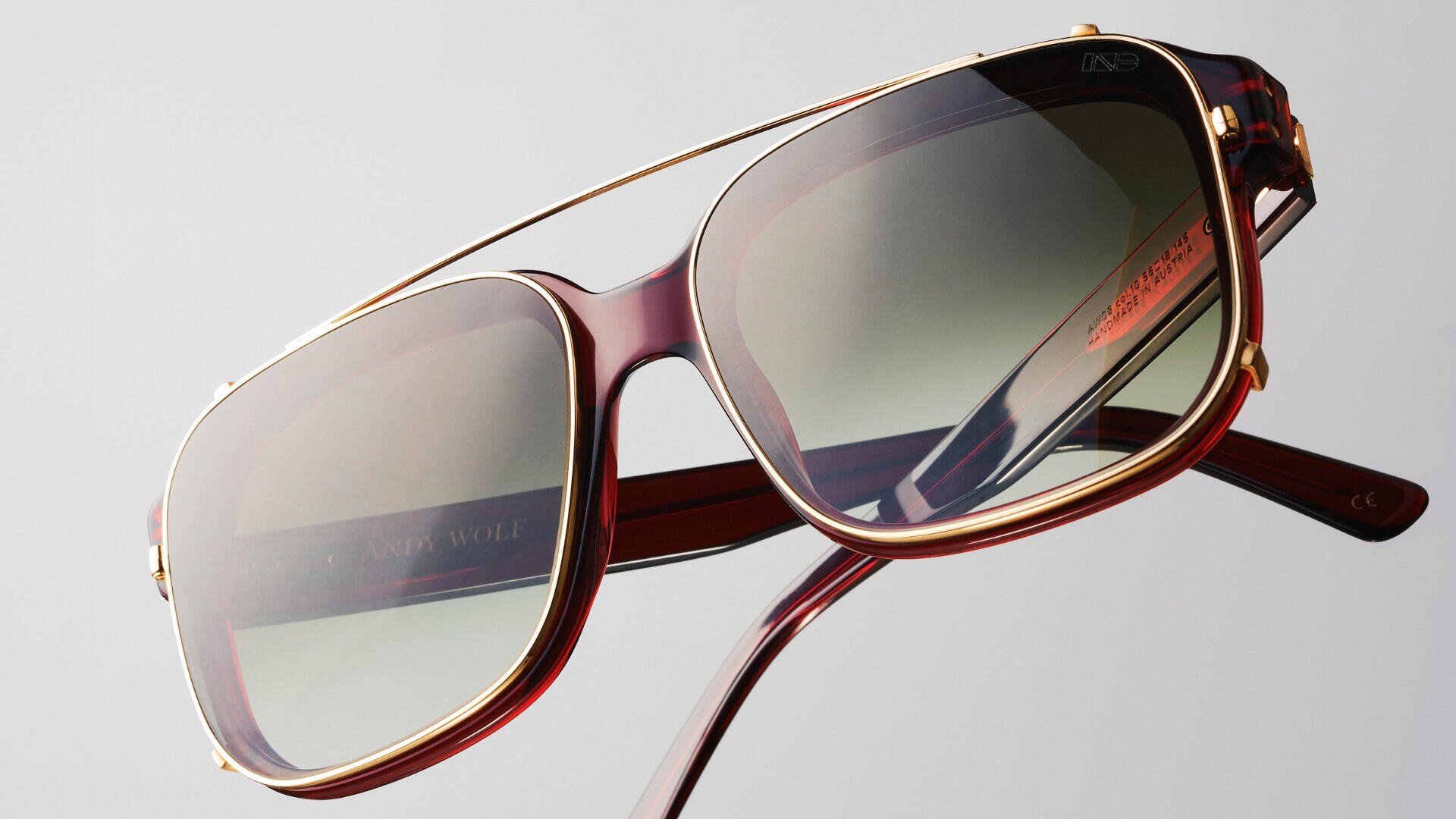 Nahaufnahme einer stylischen Sonnenbrille mit gold-rotem Rahmen, großen, getönten Gläsern und einem dünnen Goldbalken am oberen Rand vor einem schlichten hellgrauen Hintergrund.