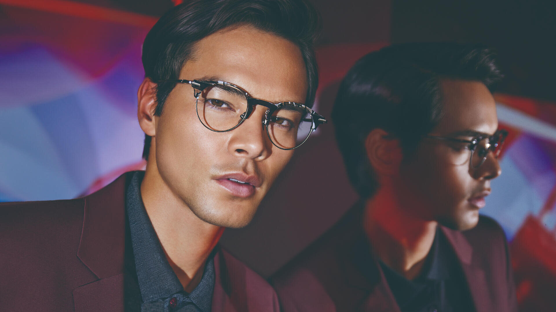 Ein Mann mit runder Brille und kastanienbrauner Jacke blickt mit ernstem Gesichtsausdruck in die Kamera, während sein Spiegelbild neben ihm vor einem bunten, unscharfen Hintergrund erscheint.