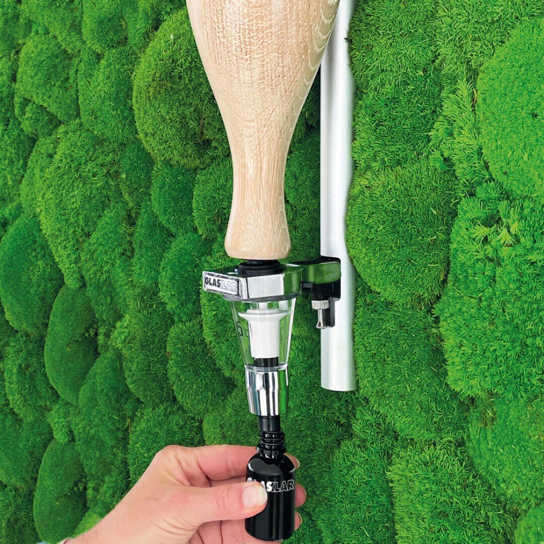 Eine Hand gibt mithilfe eines Spenders aus Metall und Kunststoff eine Flüssigkeit aus einem umgedrehten hölzernen Baseballschläger, der an einer grünen Mooswand befestigt ist, in eine kleine schwarze Flasche.