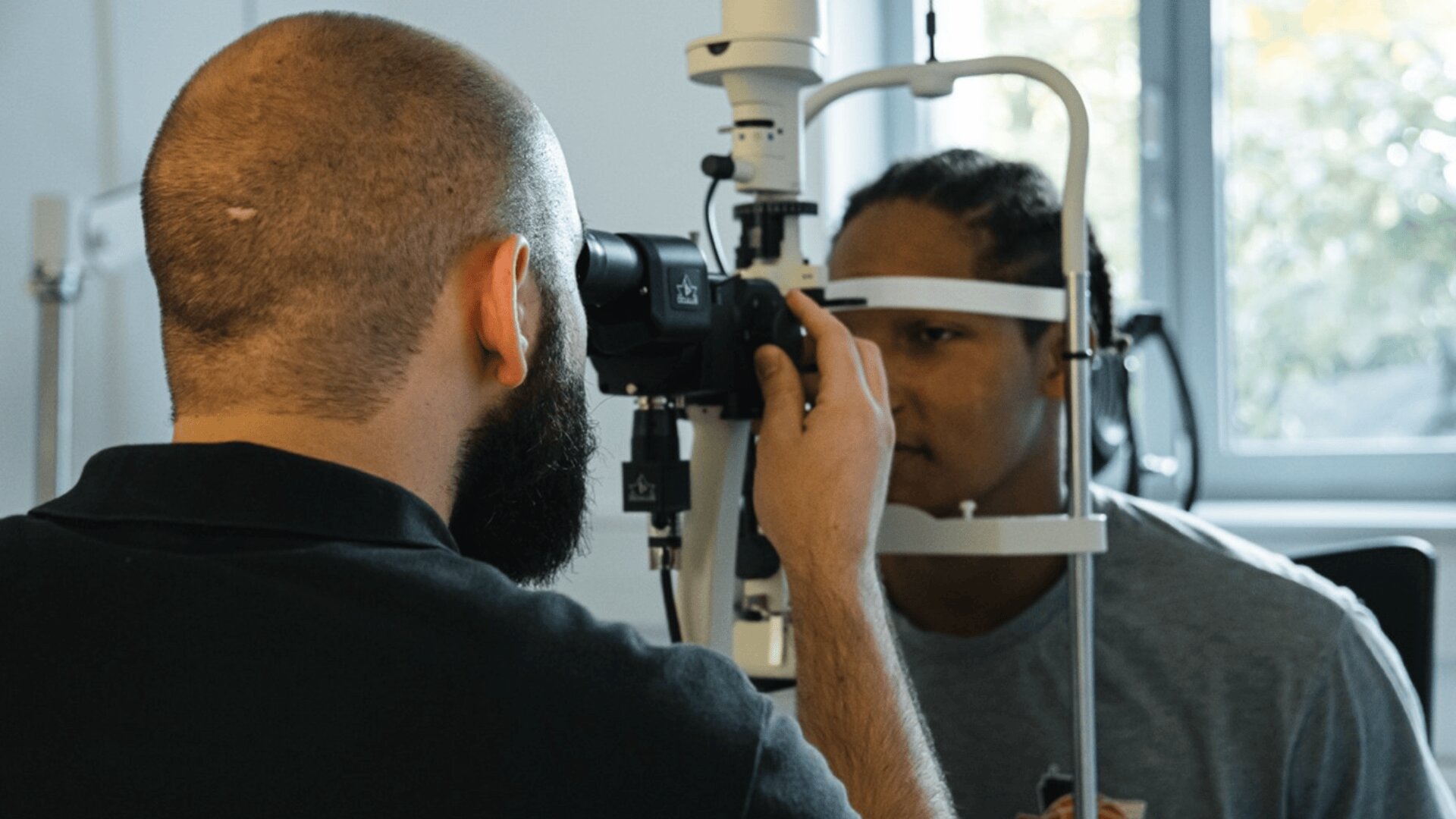 Ein Augenarzt untersucht die Augen eines Patienten mit einer Spaltlampe in einem gut beleuchteten Raum, wobei der Patient sitzt und in das Gerät schaut.