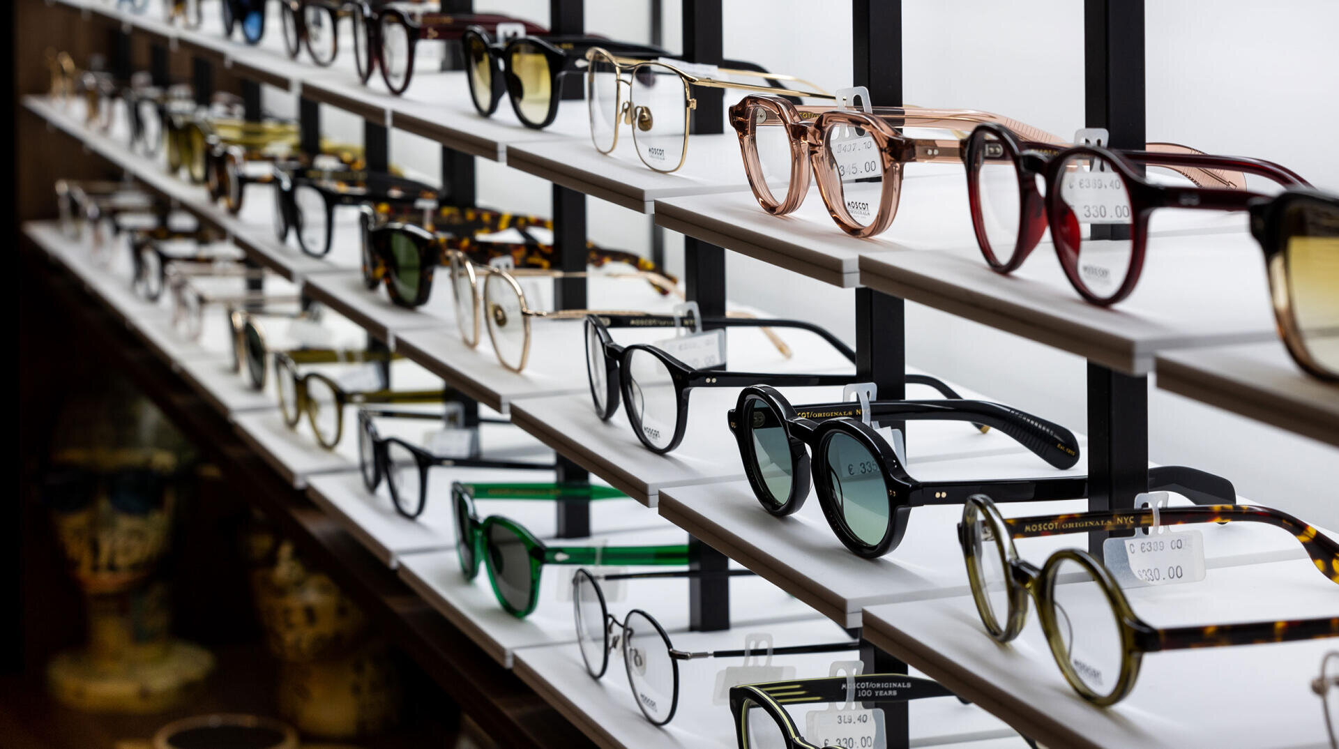 Reihen von Brillen und Sonnenbrillen in verschiedenen Formen, Farben und Stilen, die in einem Brillengeschäft übersichtlich in weißen Regalen ausgestellt sind. Preisschilder hängen an den Rahmen, und der Hintergrund ist sanft beleuchtet.