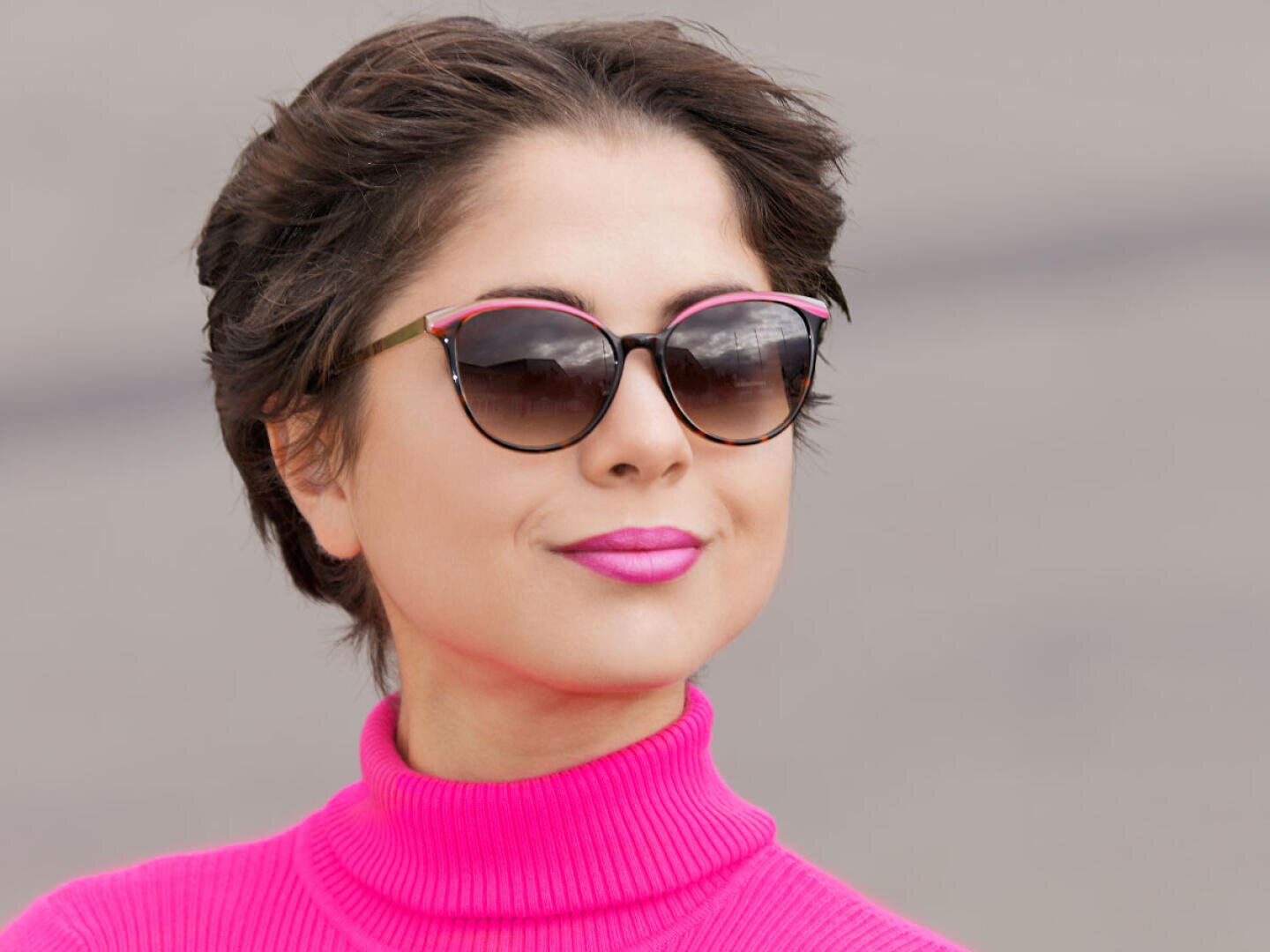 A woman wearing a light pink turtleneck sweater and sunglasses with cat-eye frames stands outdoors, smiling confidently with pink lipstick. The background is blurred and neutral.