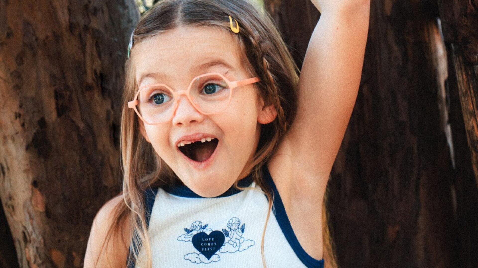 A young child with long brown hair, wearing light pink glasses and a white tank top, smiles excitedly with one arm raised, standing in front of large tree trunks.
