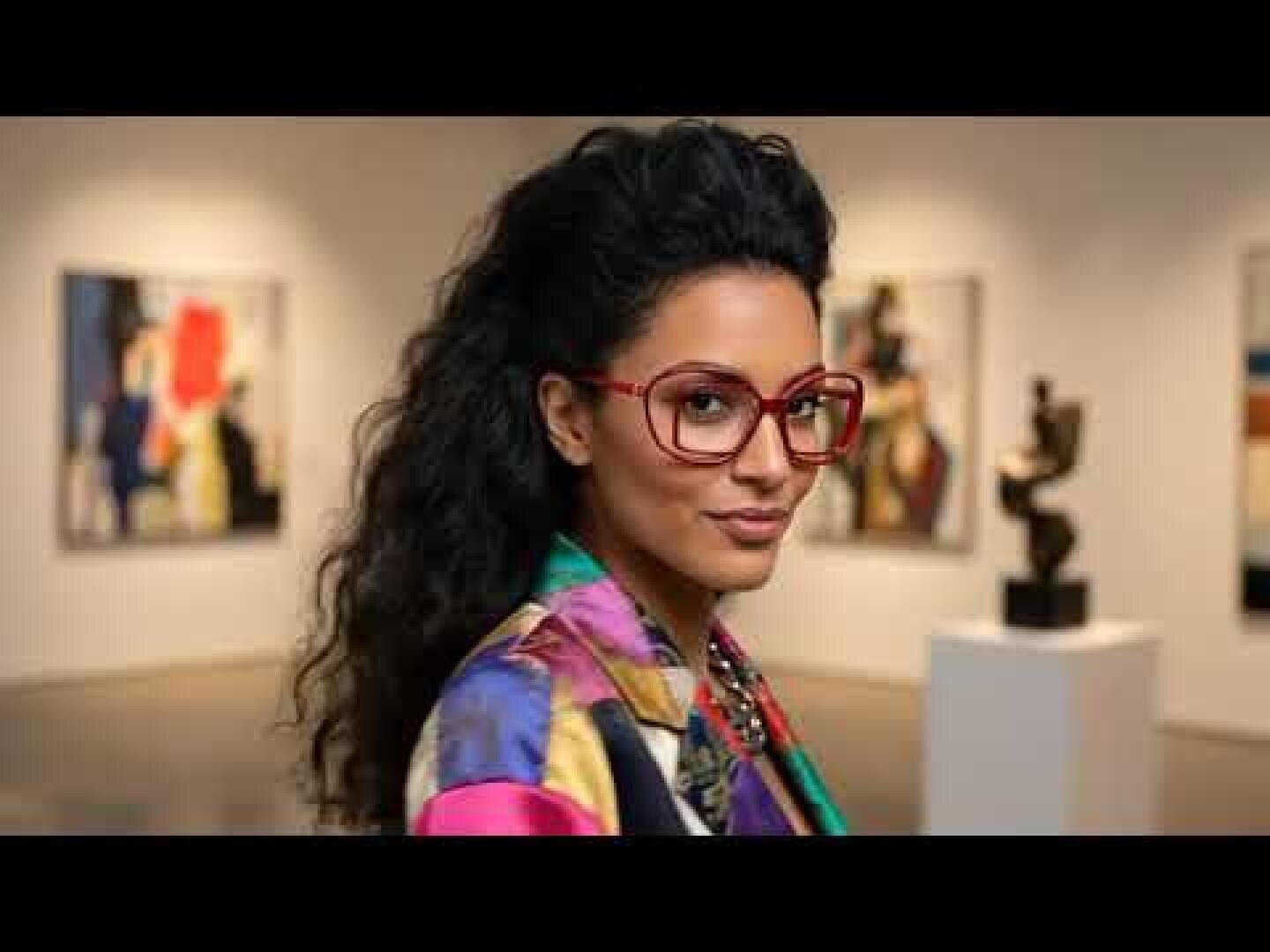 A woman with long curly hair and large red glasses stands in an art gallery wearing a colorful patterned jacket. Abstract paintings and a sculpture can be seen in the background.