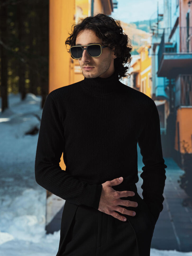 A man with dark sunglasses and a black turtleneck sweater stands outside, one hand on his waist. Snow and colorful buildings can be seen in the background.