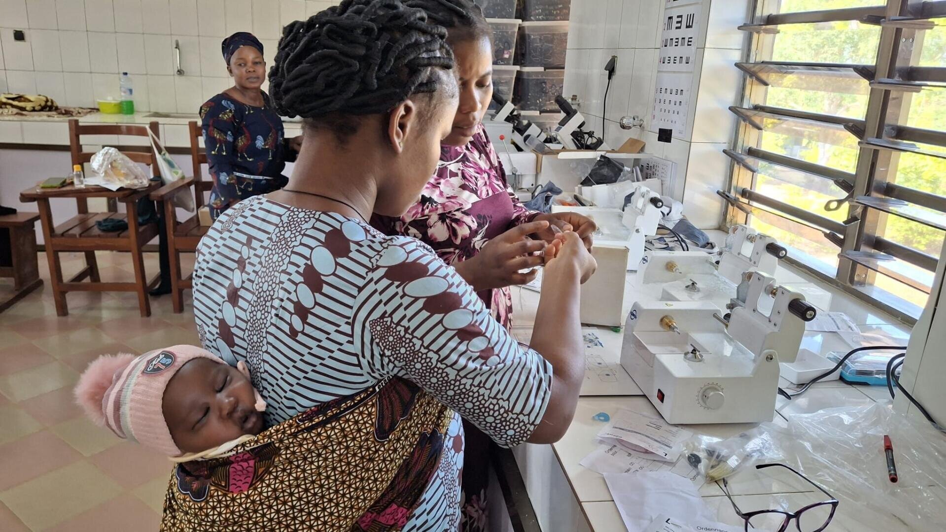Drei Frauen arbeiten gemeinsam in einem Labor mit medizinischen Geräten; eine von ihnen hat ein Baby auf den Rücken gewickelt. Im Hintergrund sind weitere Frauen zu sehen, und durch große Fenster strömt Sonnenlicht herein.