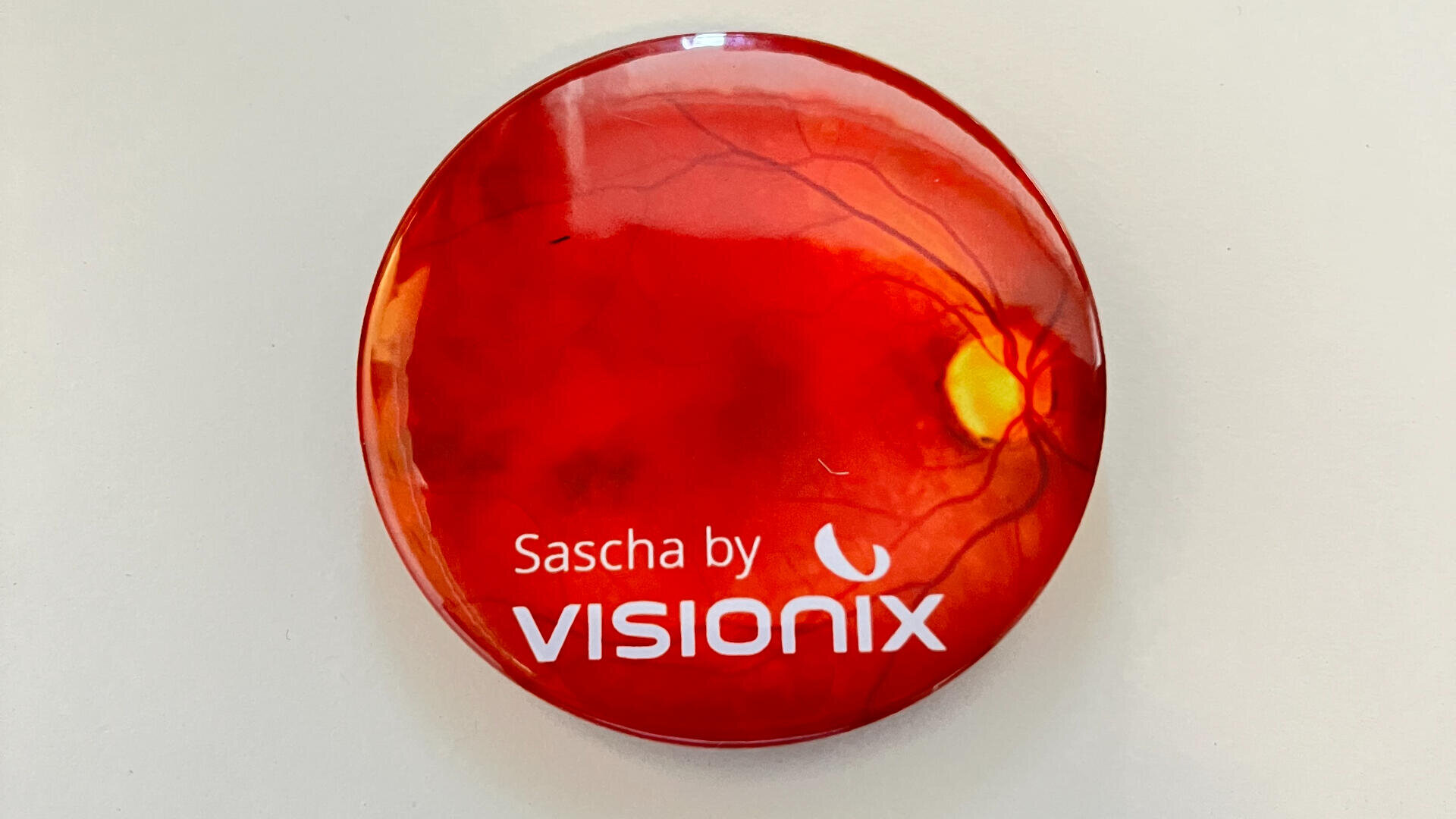Ein runder, roter Button zeigt auf der Vorderseite ein Bild, das an die Innenseite eines Auges erinnert, und den Text „Sascha by VISIONIX“ in weißen Buchstaben.