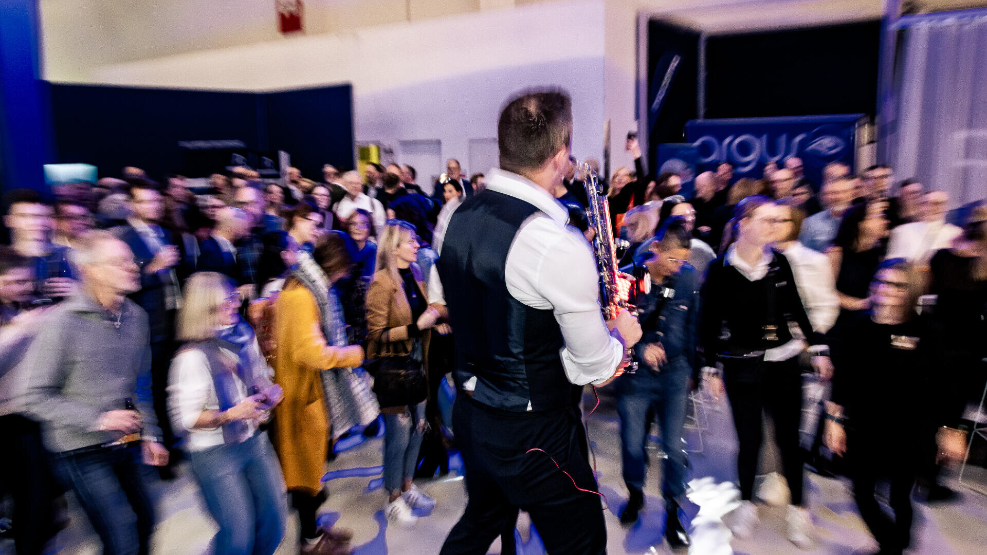Ein Musiker in einer Weste spielt Saxophon, während er bei einer Indoor-Veranstaltung durch eine lebhafte Menge geht; viele Leute schauen zu und machen Fotos.