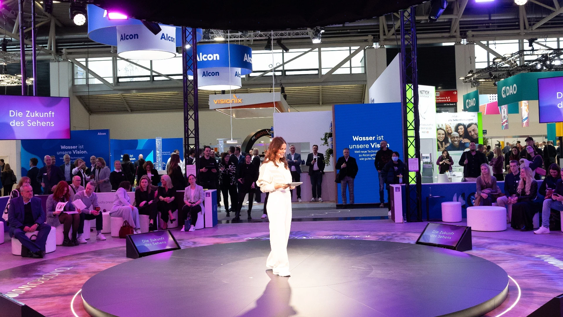 A woman in white stands on a round stage, speaking to an audience at a tech or science expo. Booths and digital displays with German text and company logos are visible in the background.