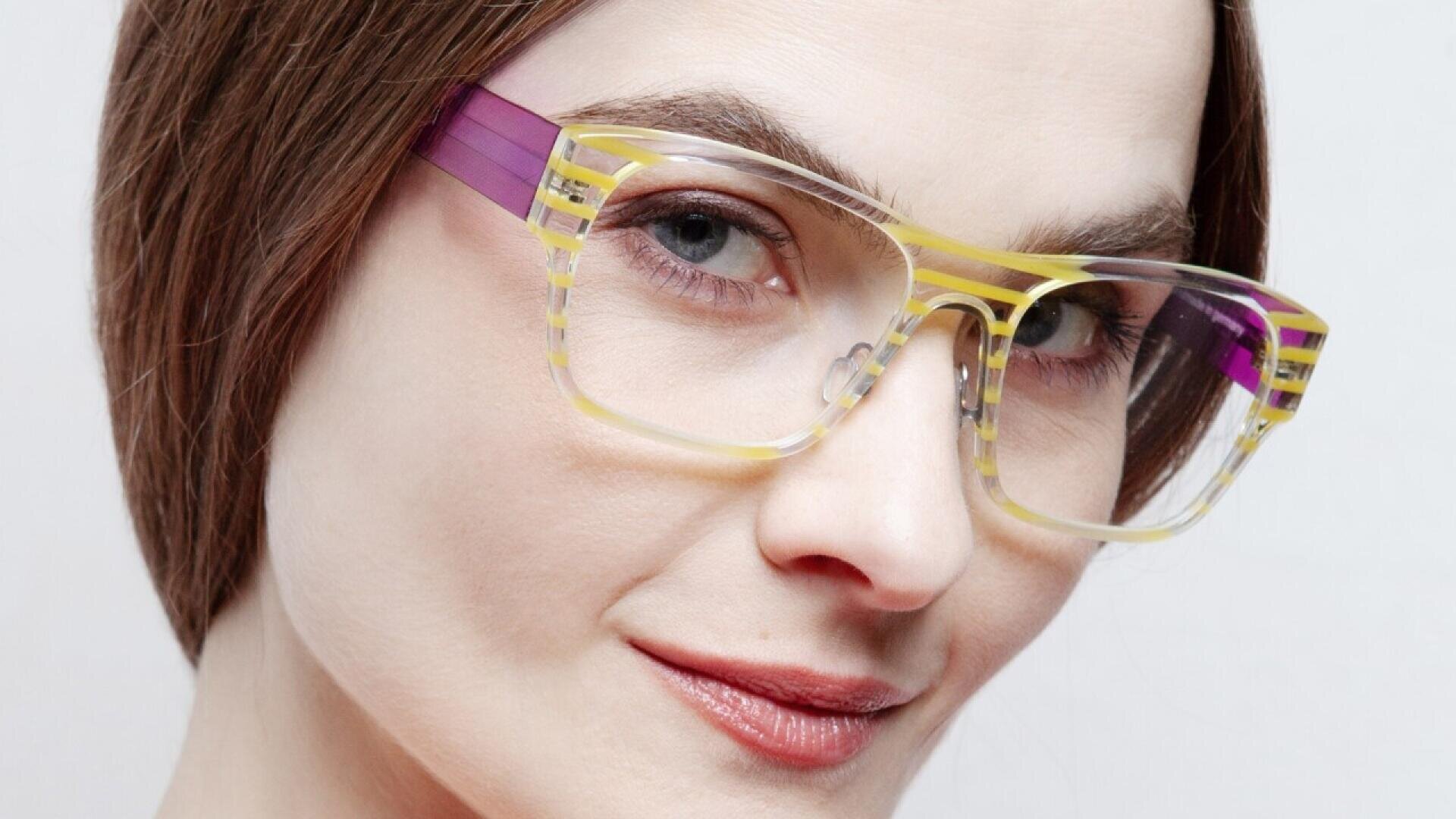 A woman with fair skin and straight brown hair wears clear eyeglasses with yellow and purple accents, looking confidently at the camera with a slight smile.