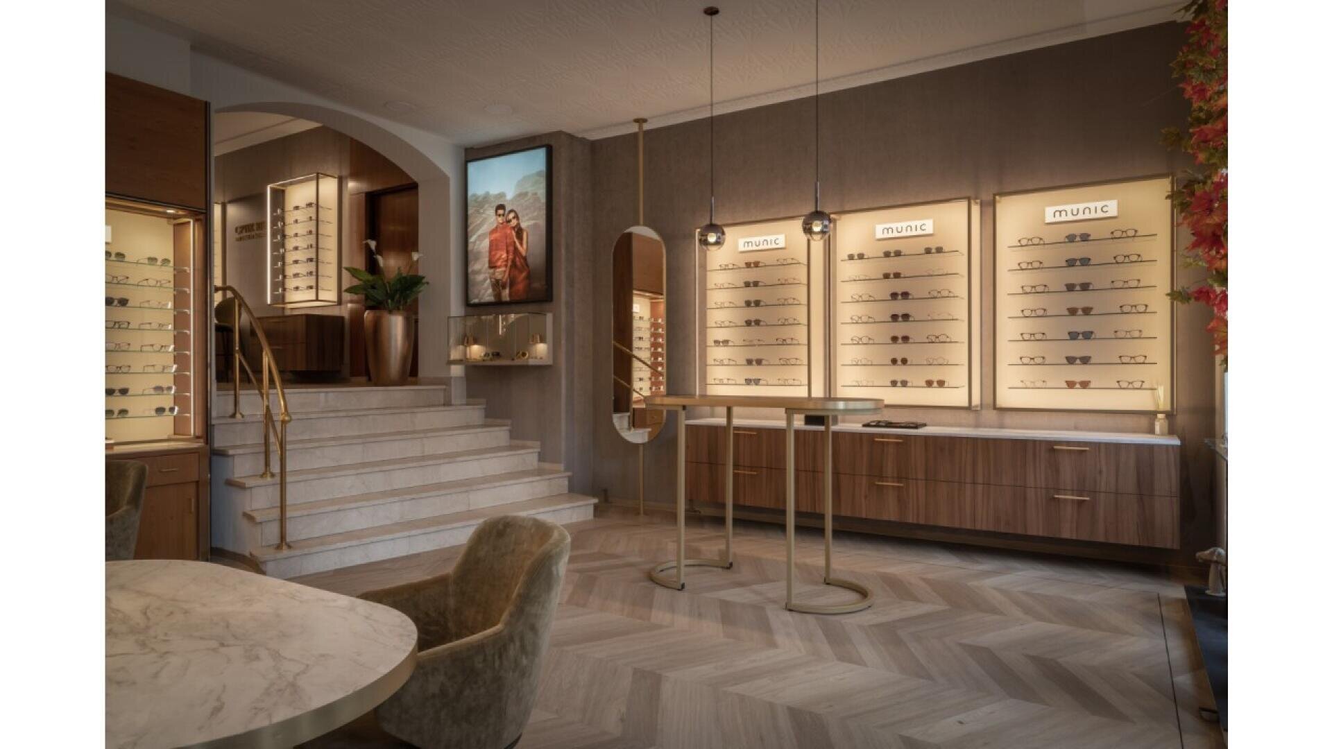 A stylish eyewear store features illuminated display walls with glasses, a marble table with chairs, wood cabinets, a staircase with a gold handrail, and warm, modern lighting.