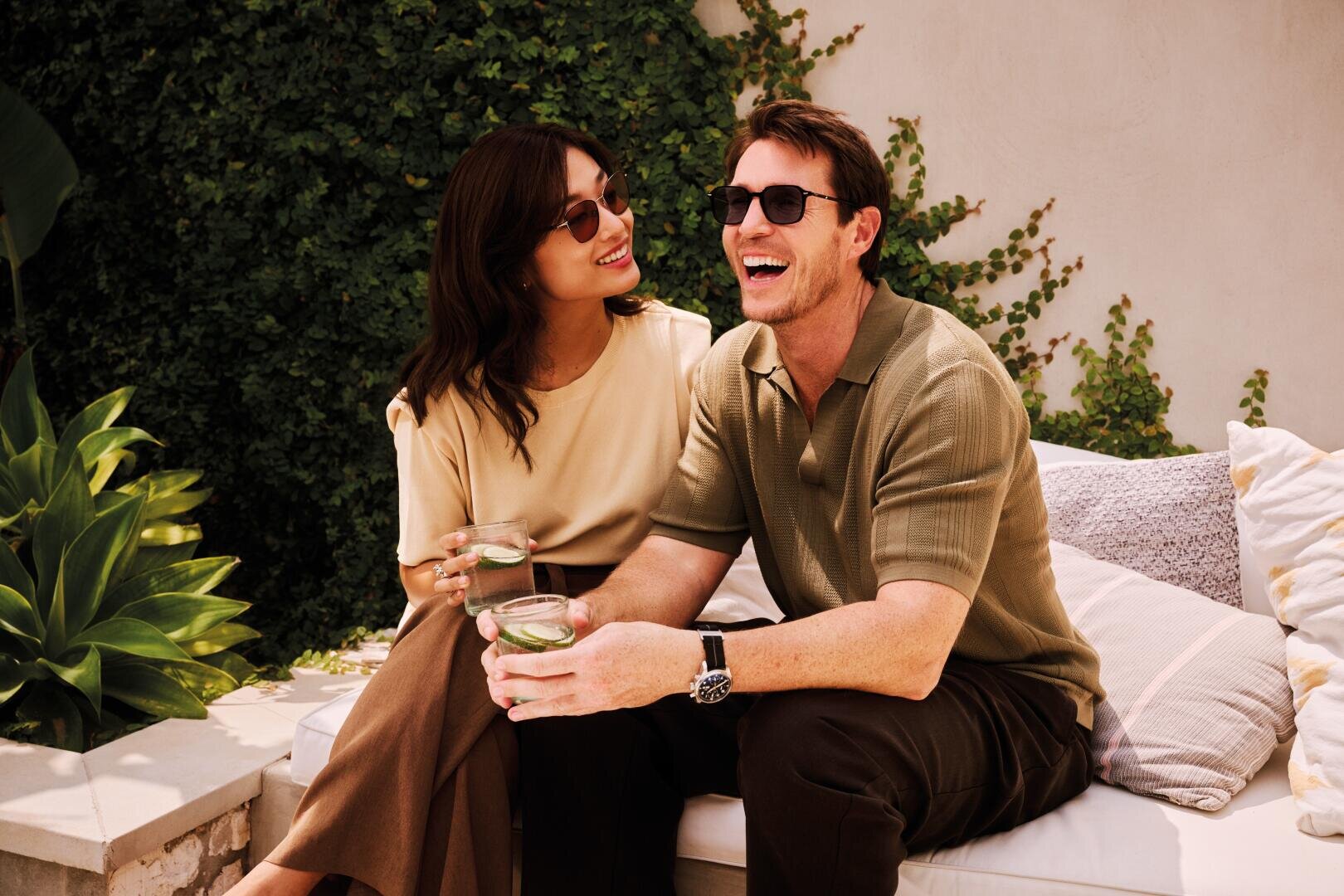 Ein lächelnder Mann und eine Frau mit Sonnenbrille sitzen auf einer Couch im Freien, umgeben von Grün, halten Getränke in der Hand und genießen einen entspannten Moment in der Sonne.