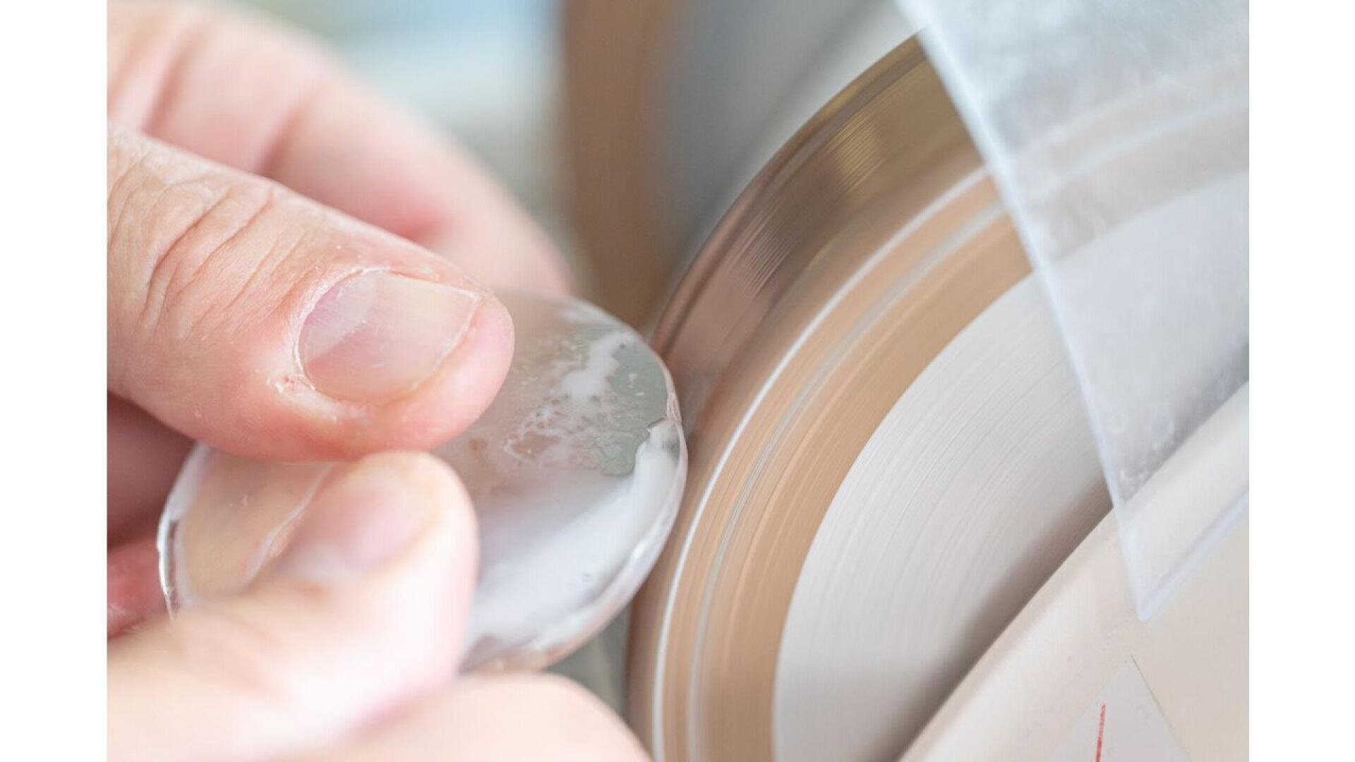 A close-up of hands holding a lens or piece of glass against a rotating polishing or grinding wheel, showing the process of shaping or smoothing the material.