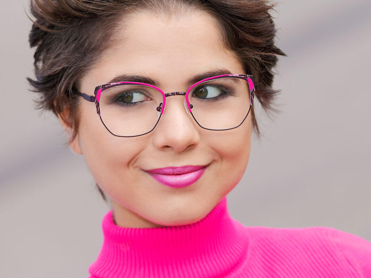 A person with short brown hair wears large, elegant glasses and a light pink turtleneck sweater and smiles slightly. Her lipstick matches the color of her sweater.
