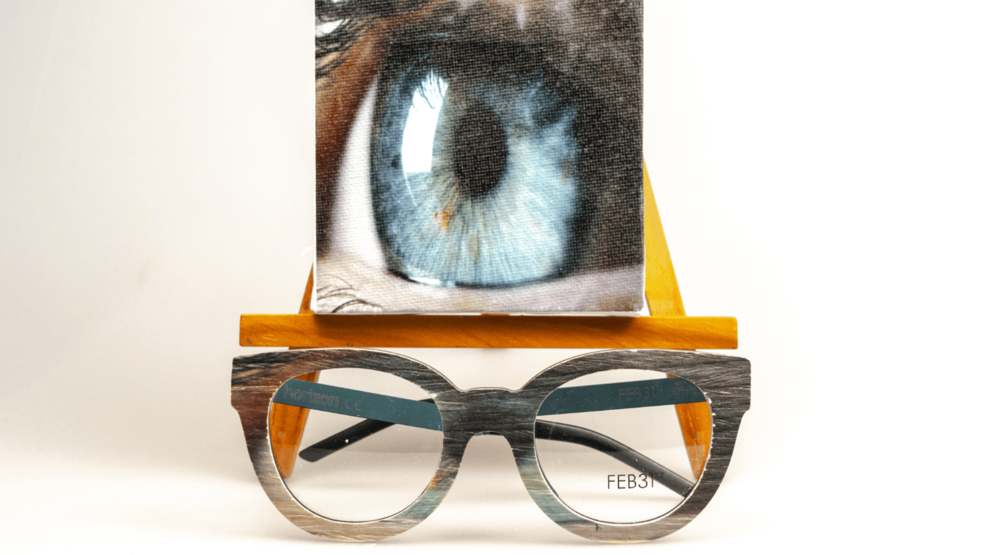 Vor einer kleinen Staffelei, auf der ein blaues Auge in Großaufnahme abgebildet ist, steht eine grau umrandete Brille, die den Eindruck erweckt, als würde das Auge durch die Brille schauen.