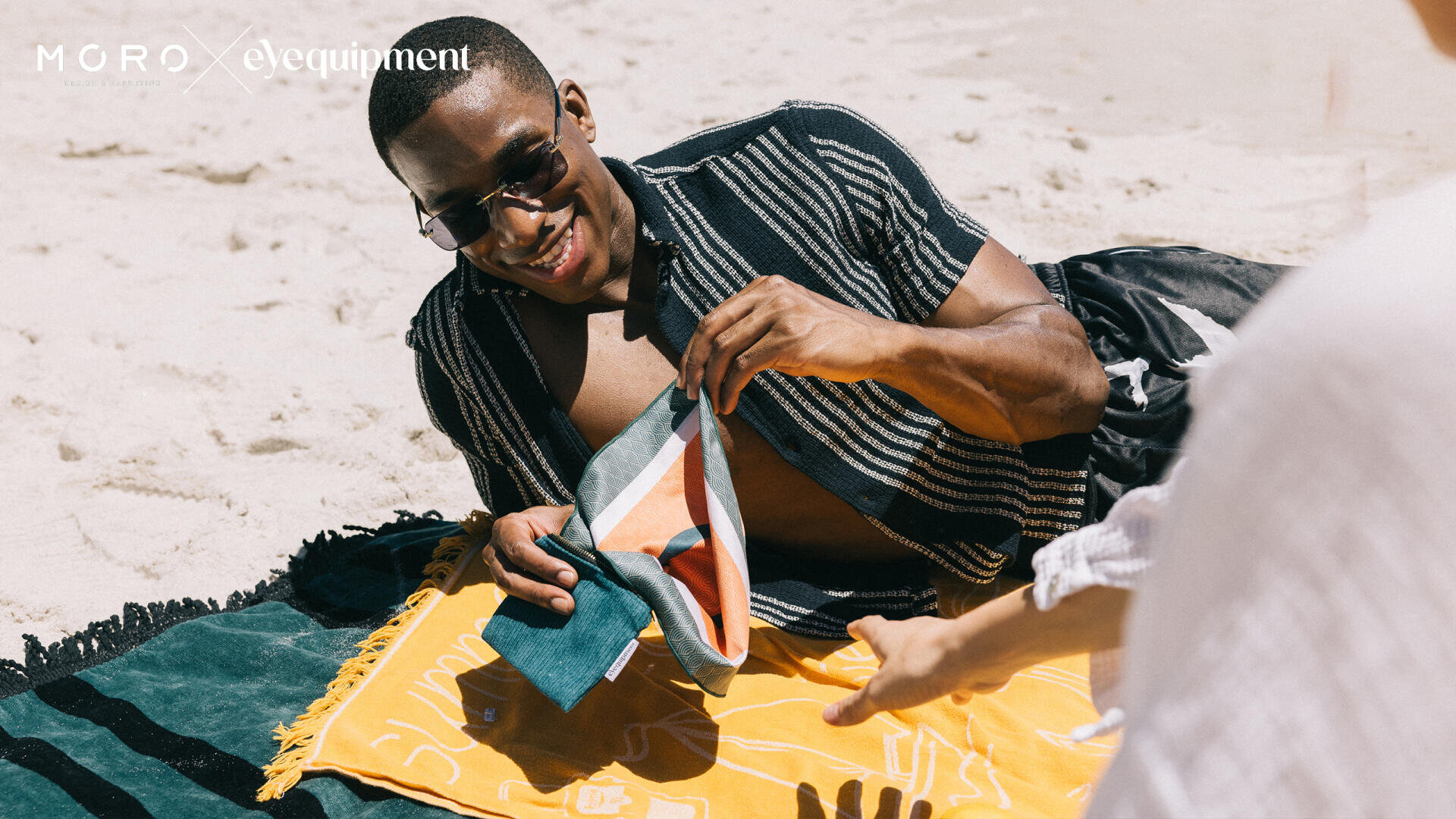 Ein Mann mit Sonnenbrille lächelt, während er auf einem Strandtuch liegt und einen bunten Beutel in der Hand hält. Er liegt im Sand, der Arm einer anderen Person streckt sich nach ihm aus. Die Szene wirkt entspannt und sonnig.