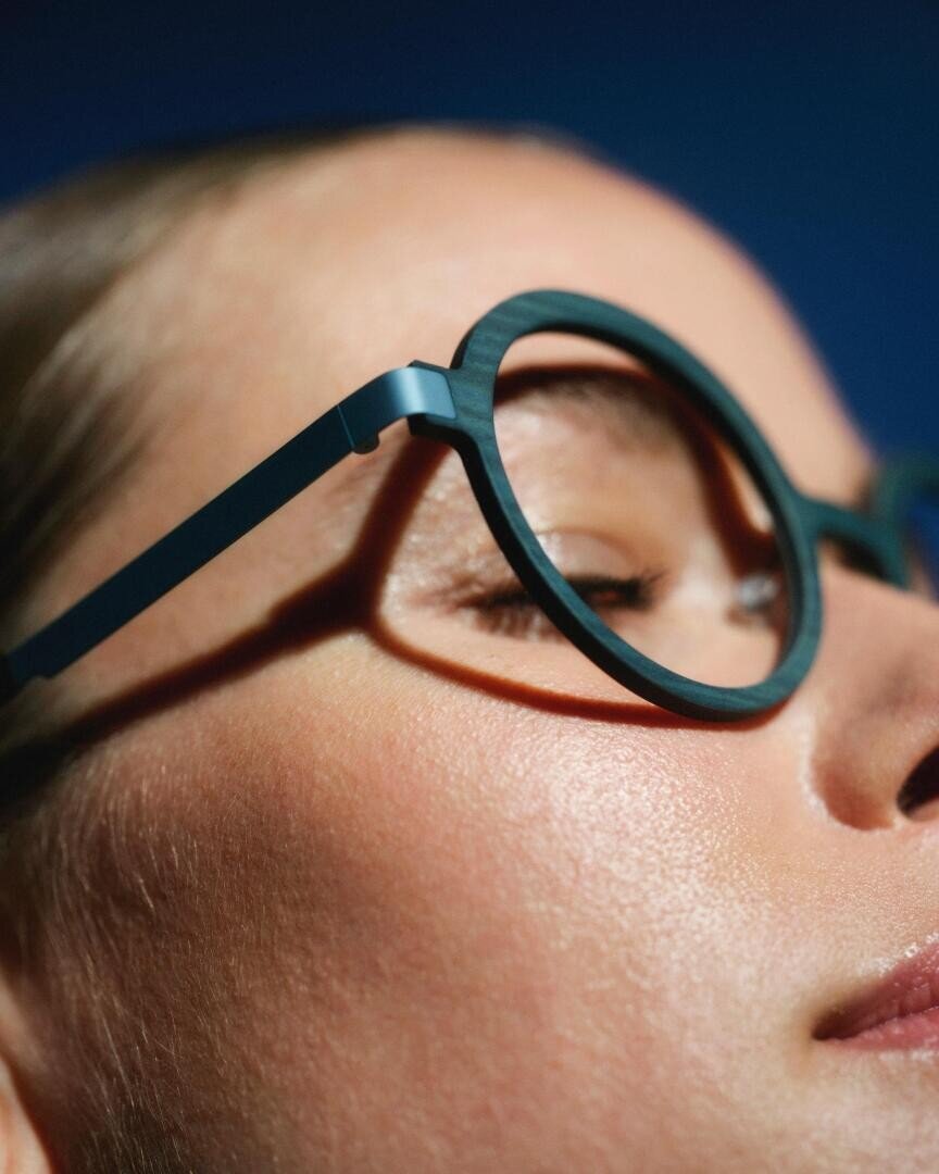 Close-up of a persons face wearing round, teal eyeglasses with eyes closed, showing smooth skin and a calm expression against a deep blue background.