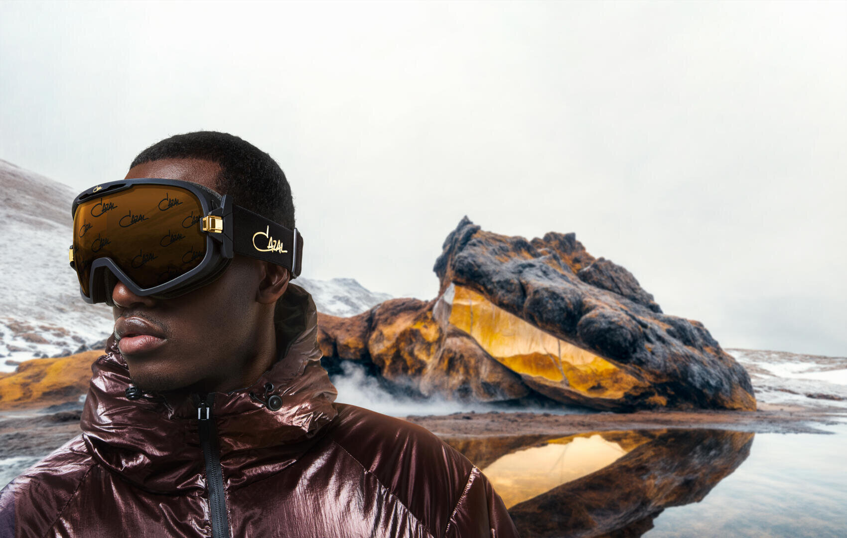 Eine Person mit reflektierender Skibrille und glänzender brauner Jacke steht vor einer felsigen, schneebedeckten Landschaft mit aufsteigendem Dampf und einer gelb leuchtenden Höhle, die sich im stillen Wasser spiegelt.