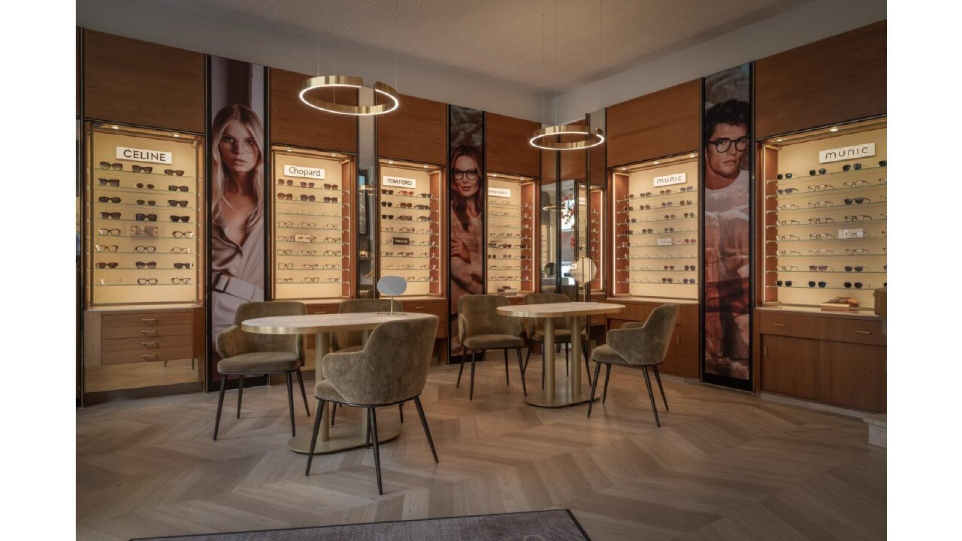 A stylish eyewear boutique with wooden display cases showcasing various glasses. Four chairs and two round tables sit in the center, with large model photos and brand names like Celine and Munic on the walls.