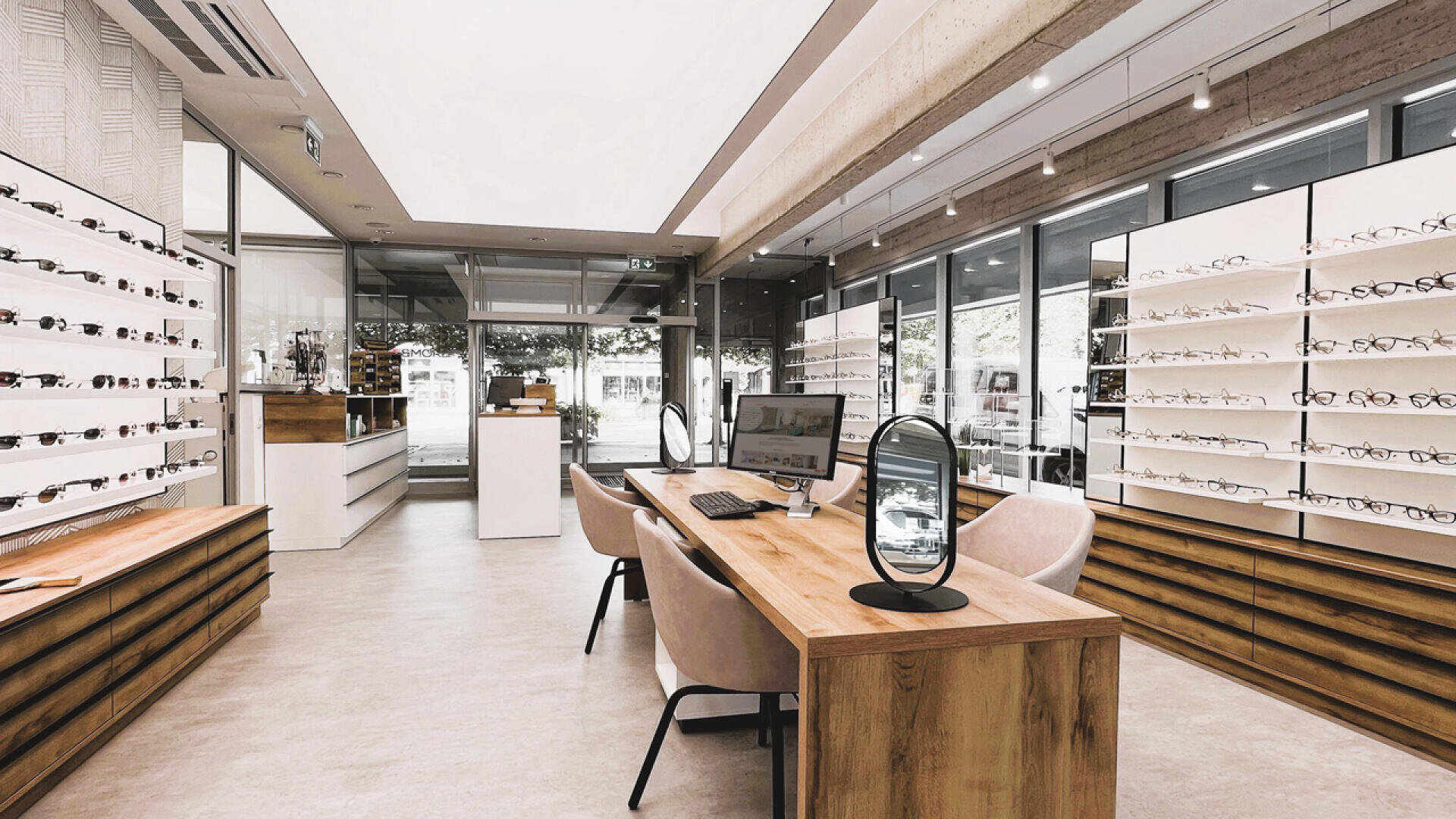 Ein modernes Optikergeschäft mit Holztischen, Regalen voller Brillen, zwei Stühlen an jedem Tisch, Computern und großen Fenstern, die natürliches Licht hereinlassen.
