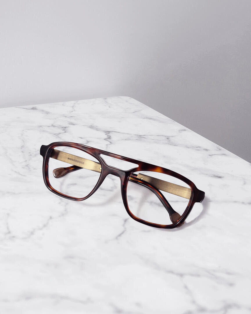 A pair of brown tortoiseshell glasses with golden temples rests on a white marble surface against a light gray background.