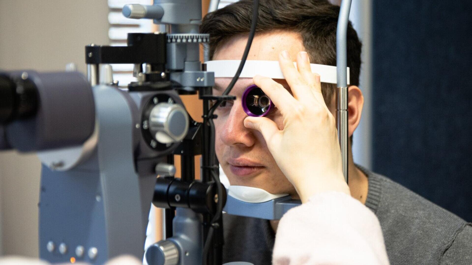 A person undergoing an eye exam, looking into a slit lamp microscope while a healthcare professional adjusts the machine to examine their eyes.